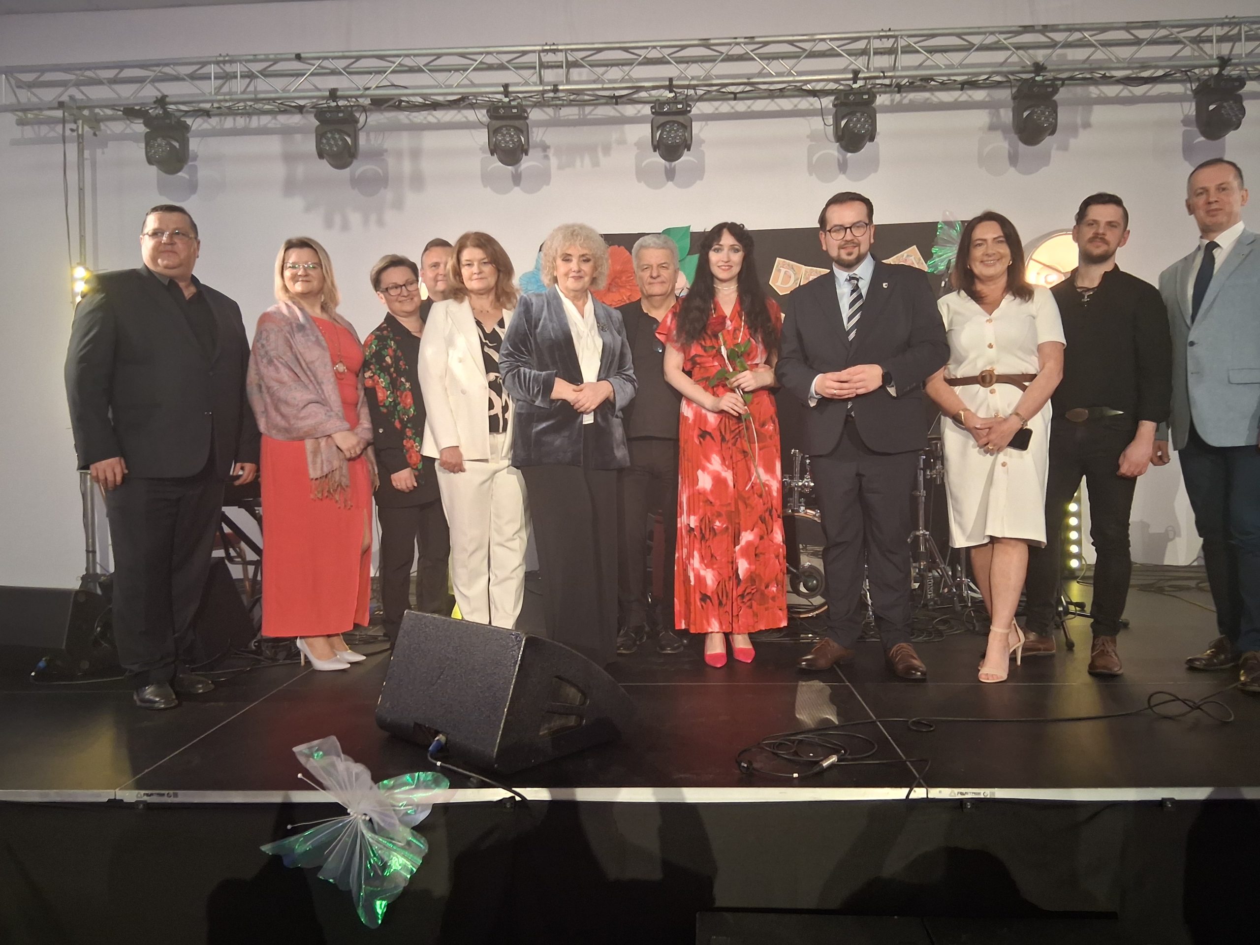 Grupa trzynastu osób stoi na scenie w eleganckich strojach. Za nimi szare tło z reflektorami, z przodu stoi kilka mikrofonów. Kobieta w centrum trzyma różę i ma czerwoną sukienkę w kwiaty.