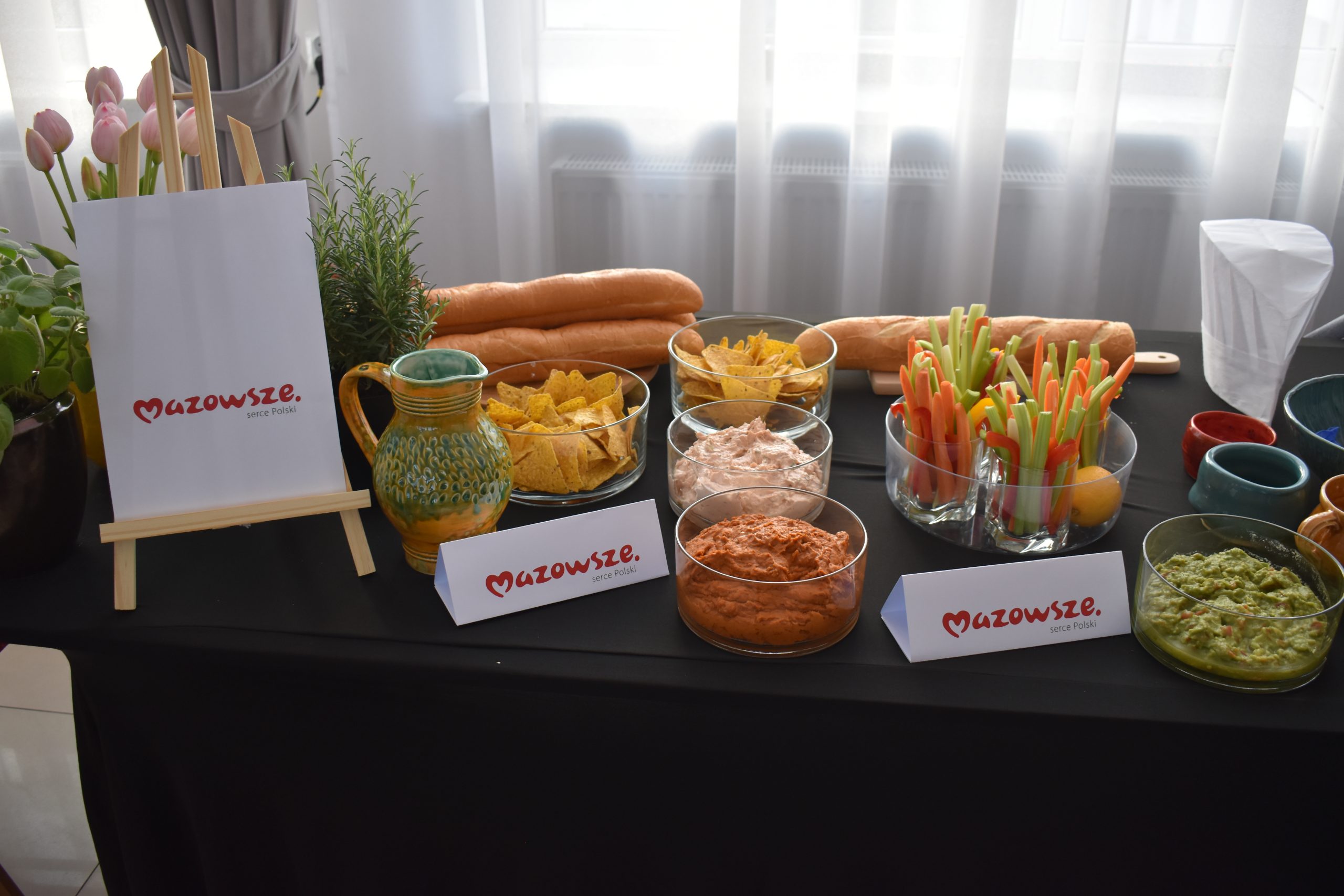 Na obrazie znajduje się stół z przekąskami: warzywa, dipy, nachosy. Dominuje ciemne tło, jasne zasłony z tyłu. Logo Mazowsze na widocznym znaku.