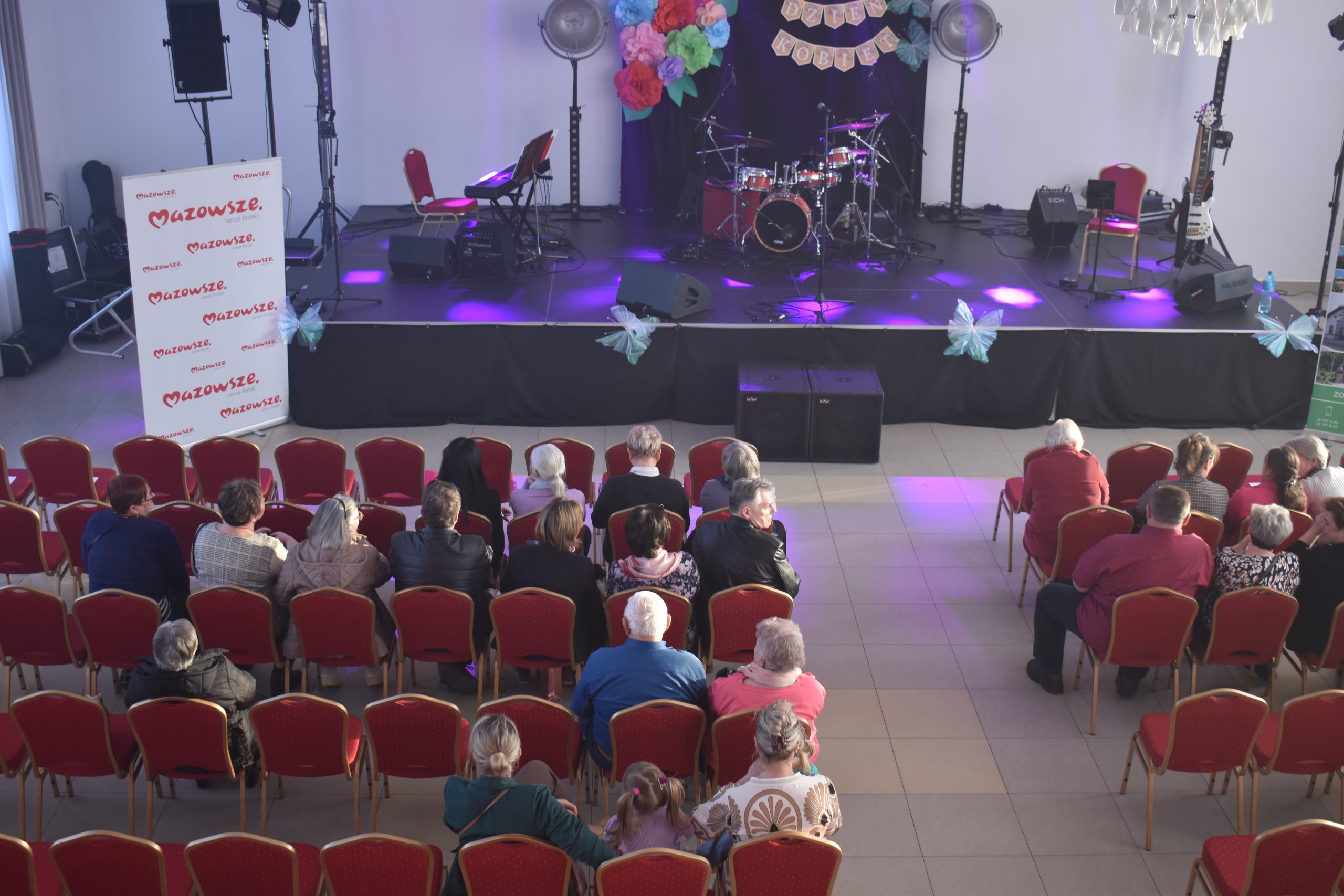 Scena z instrumentami muzycznymi i dekoracjami w tle. Przed nią siedzą ludzie na czerwonych krzesłach. Oświetlenie purpurowe i niebieskie.