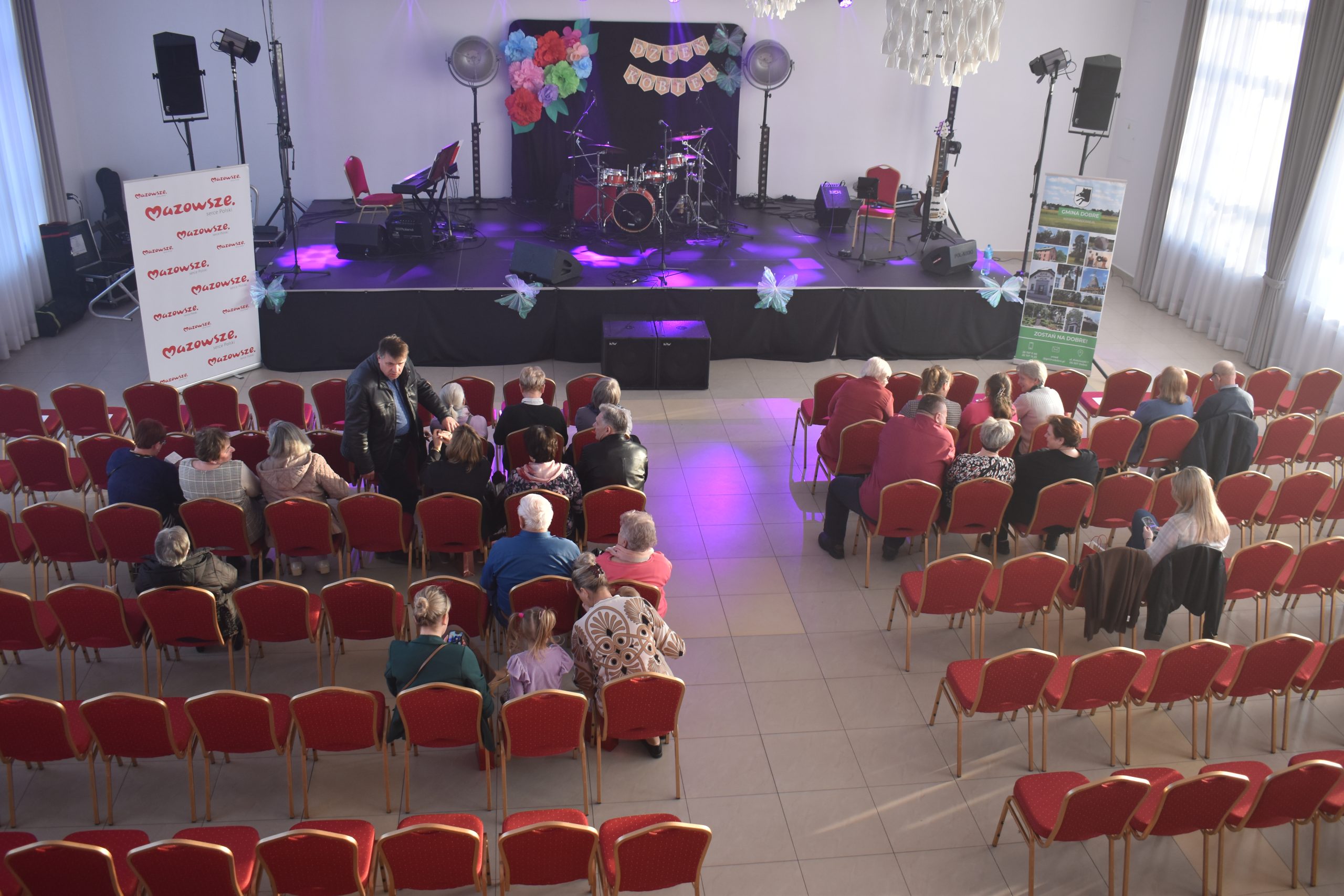 Scena udekorowana wielobarwnymi kwiatami z instrumentami muzycznymi. Przed sceną czerwone krzesła z widownią. Jasne, przestronne wnętrze z zasłonami.