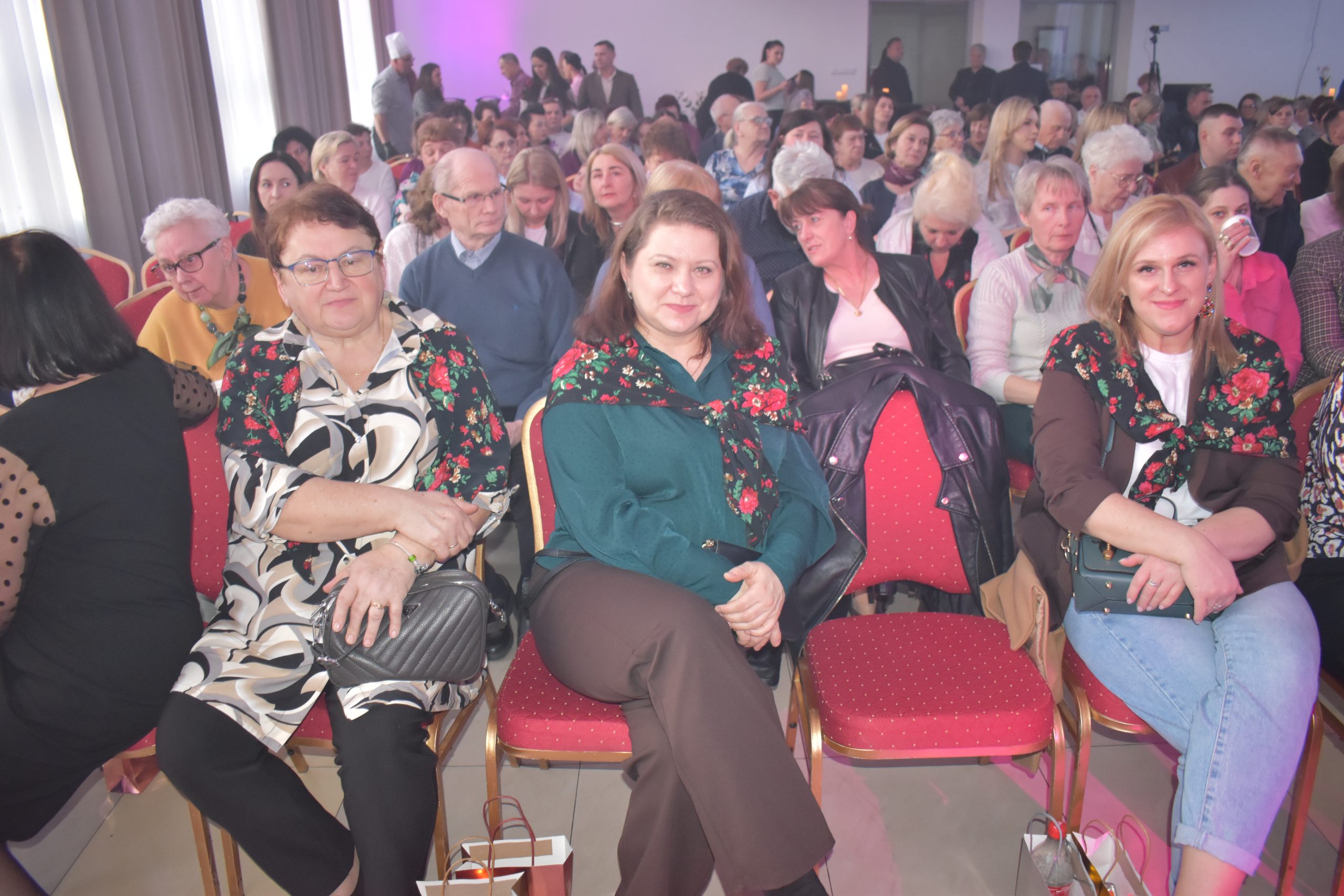 Na zdjęciu widać grupę osób siedzących na czerwonych krzesłach. Tło jest jasne z zasłonami i fioletowym światłem. Kilka osób nosi kwieciste chusty.