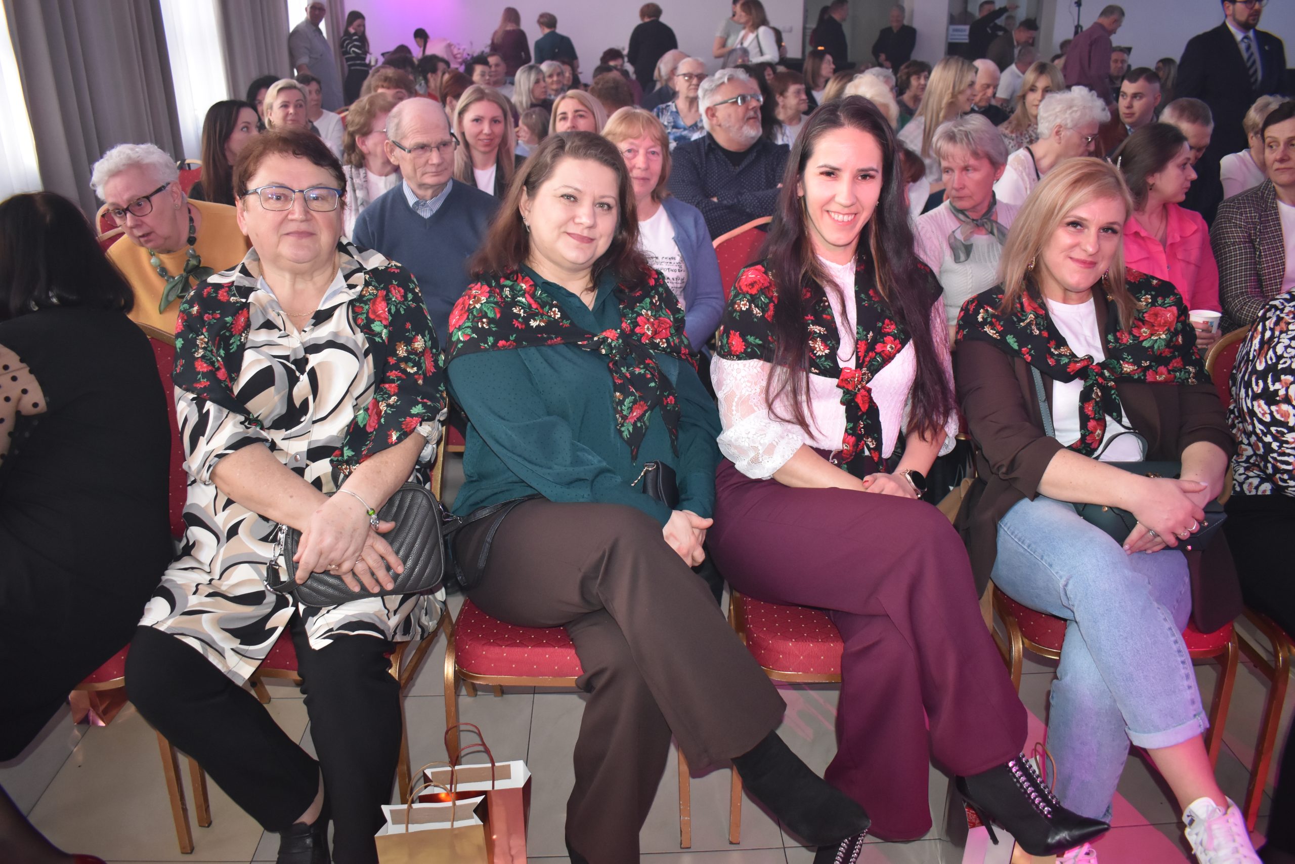 Grupa osób siedzi na krzesłach w pomieszczeniu. Kobiety na zdjęciu mają na sobie kolorowe chusty i uśmiechają się. Tło wypełnia tłum ludzi, atmosfera jest życzliwa.