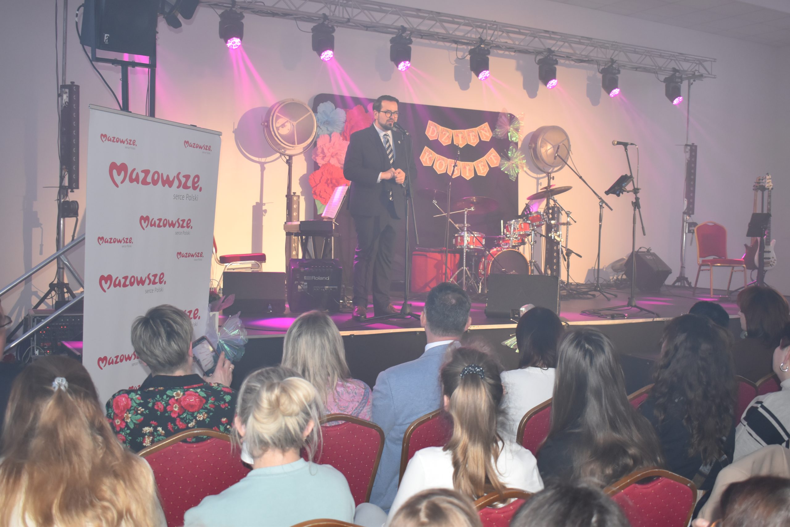 Na scenie mężczyzna w garniturze przemawia, otoczony różowym i fiołkowym światłem. W tle instrumenty muzyczne, napisy i dekoracje. Publiczność siedzi na czerwonych krzesłach, po lewej stronie biały baner z czerwonym napisem Mazowsze.