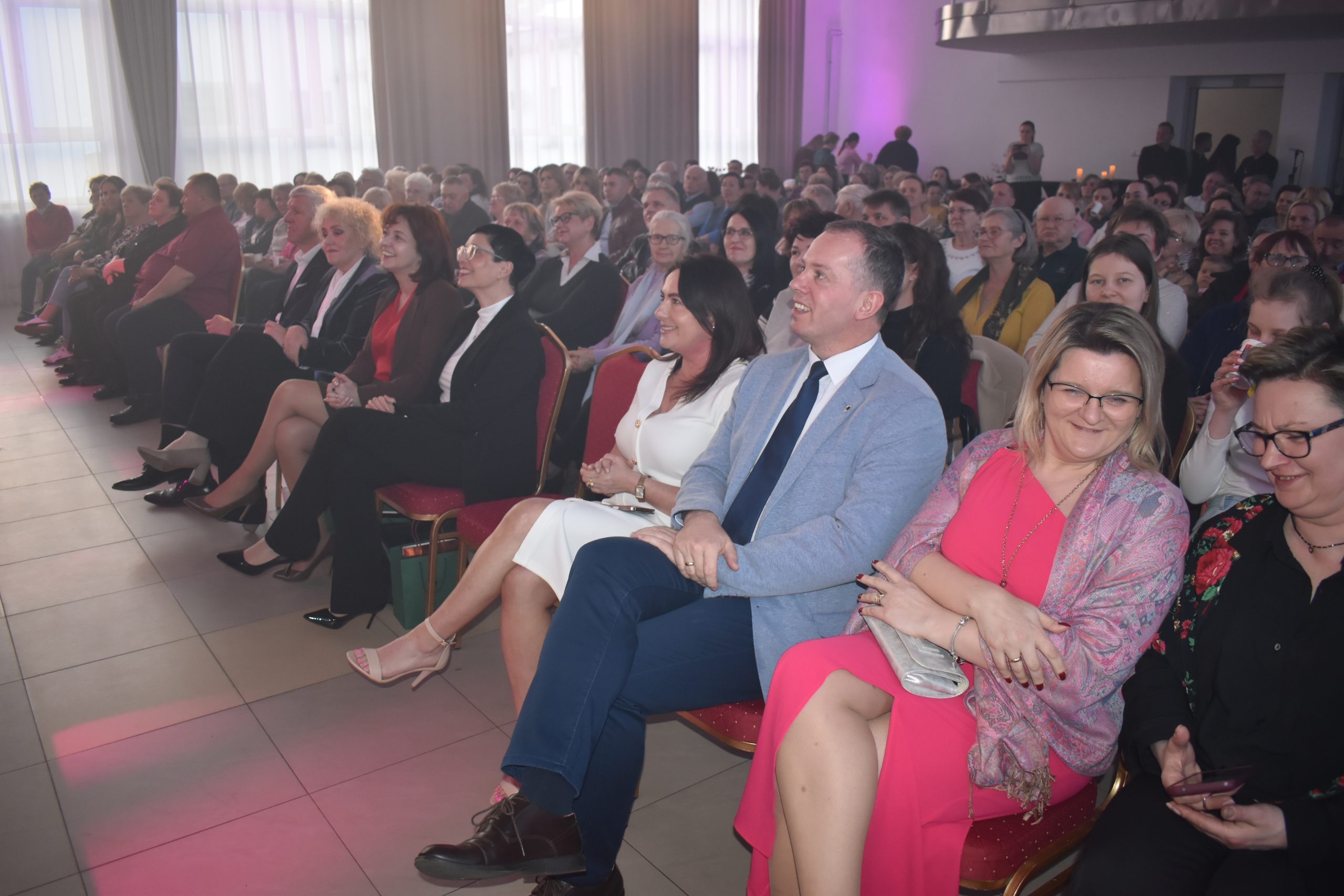 Grupa ludzi siedzi na czerwonych krzesłach, uśmiechają się w kierunku sceny. Sala jest jasna, oświetlona naturalnym i różowym światłem. W tle zasłony i duże okna.