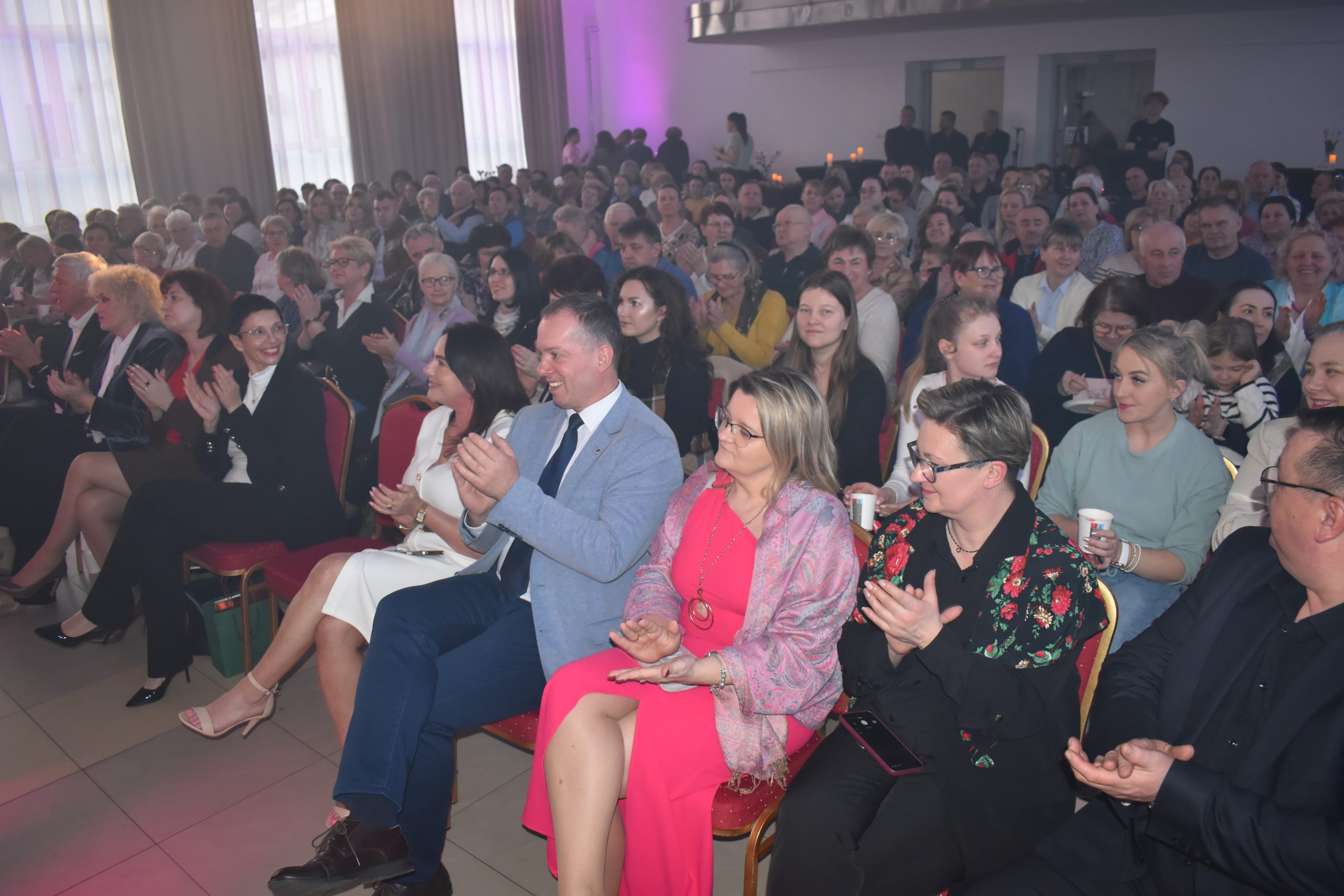 Sala pełna ludzi siedzących na czerwonych krzesłach, klaszczących. Tło jest jasne, z zasłonami i subtelnym fioletowym oświetleniem. W przodzie osoby w kolorowych strojach.