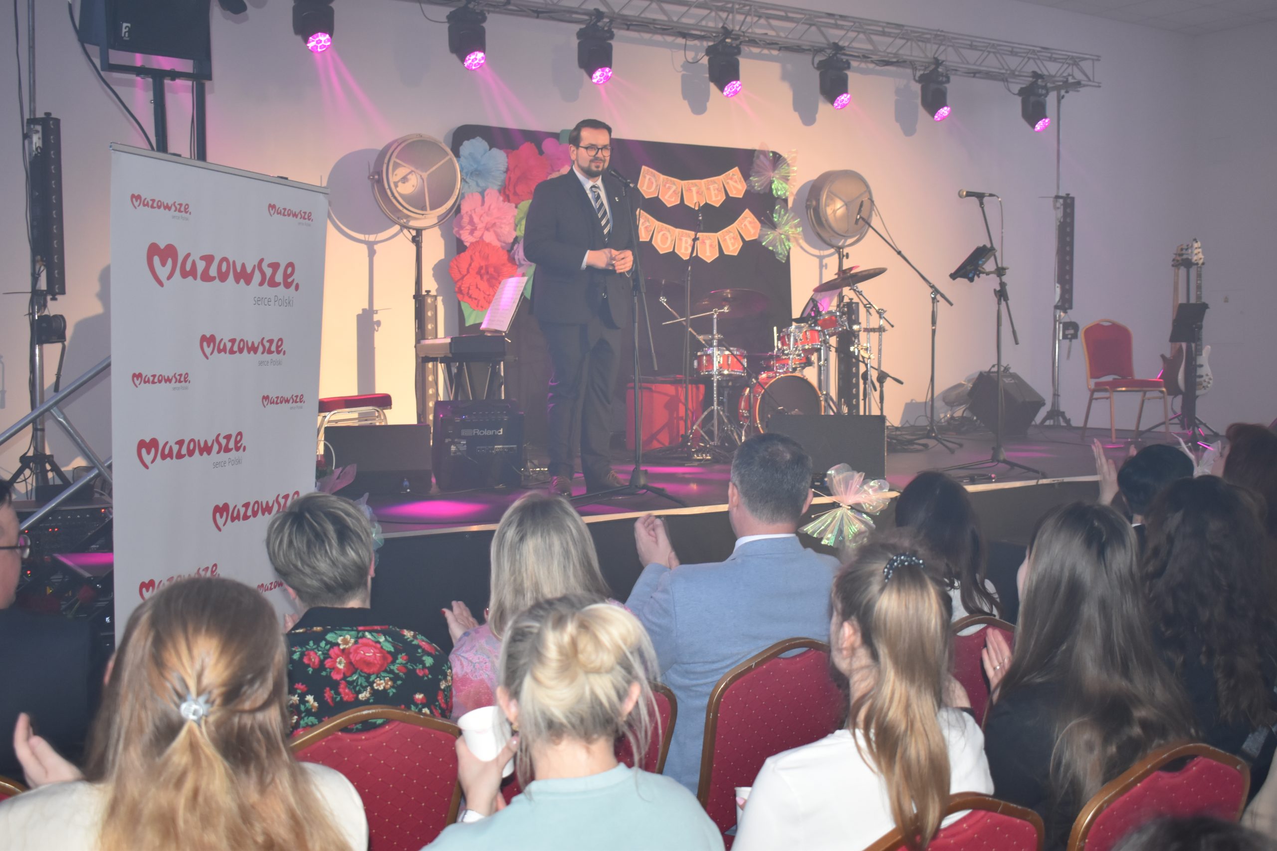 Mężczyzna w garniturze przemawia na scenie z instrumentami muzycznymi. Tło udekorowane kolorowymi kwiatami i napisem. Widzowie siedzą na czerwonych krzesłach.