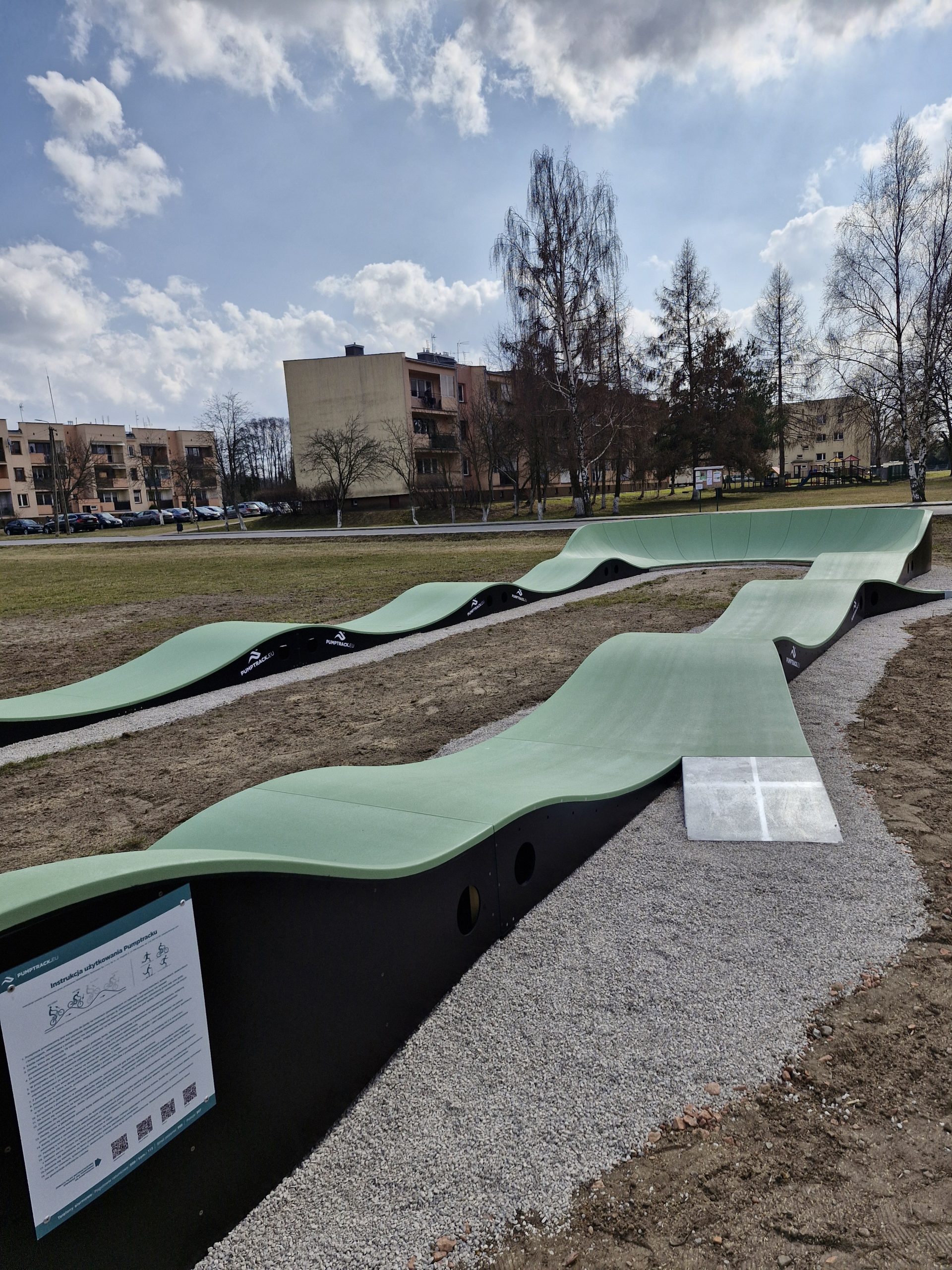 Skatepark z zielonymi falistymi rampami na tle bloków i drzew. Niebo zachmurzone, teren otoczony żwirem. Tablica informacyjna z przodu.