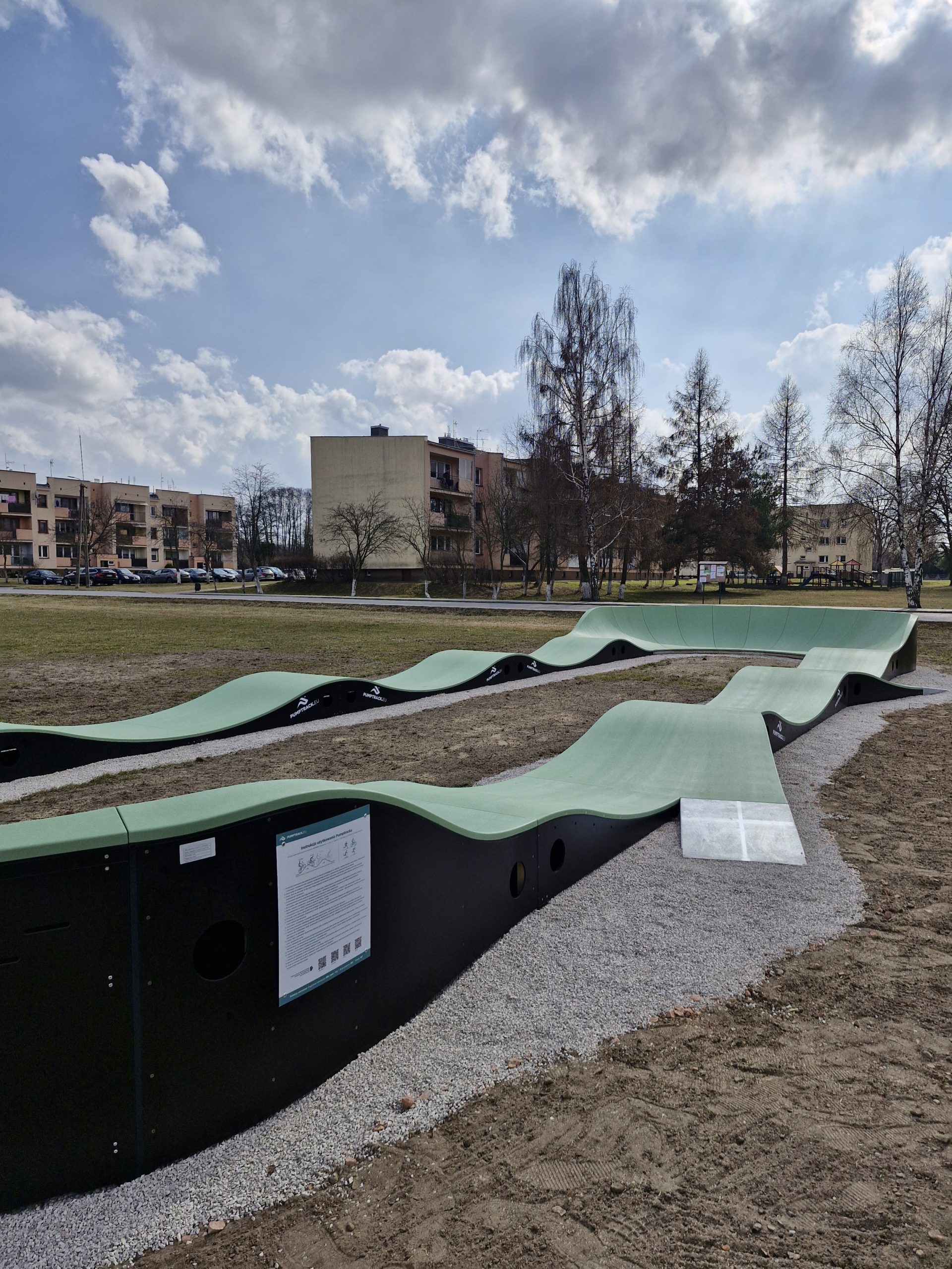 Tor rowerowy z zielonymi falistymi rampami na tle bloków i drzew. Niebo jest lekko pochmurne. Teren jest trawiasty.