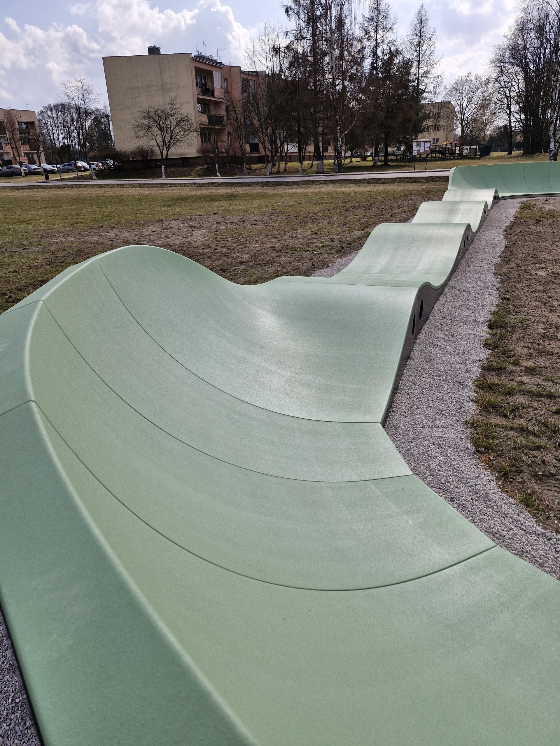 Zielony tor pumptrack na tle budynków i drzew w parku. Tło wypełniają drzewa i niskie zabudowania. Pogoda jest słoneczna z lekkim zachmurzeniem.