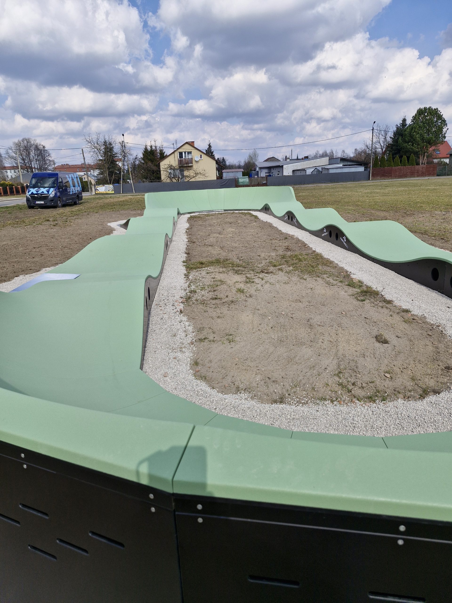 Na zdjęciu widoczny jest zielony tor pumptrack na otwartym terenie. W tle widać domy, drzewa i niebieski samochód. Na niebie znajdują się białe chmury.