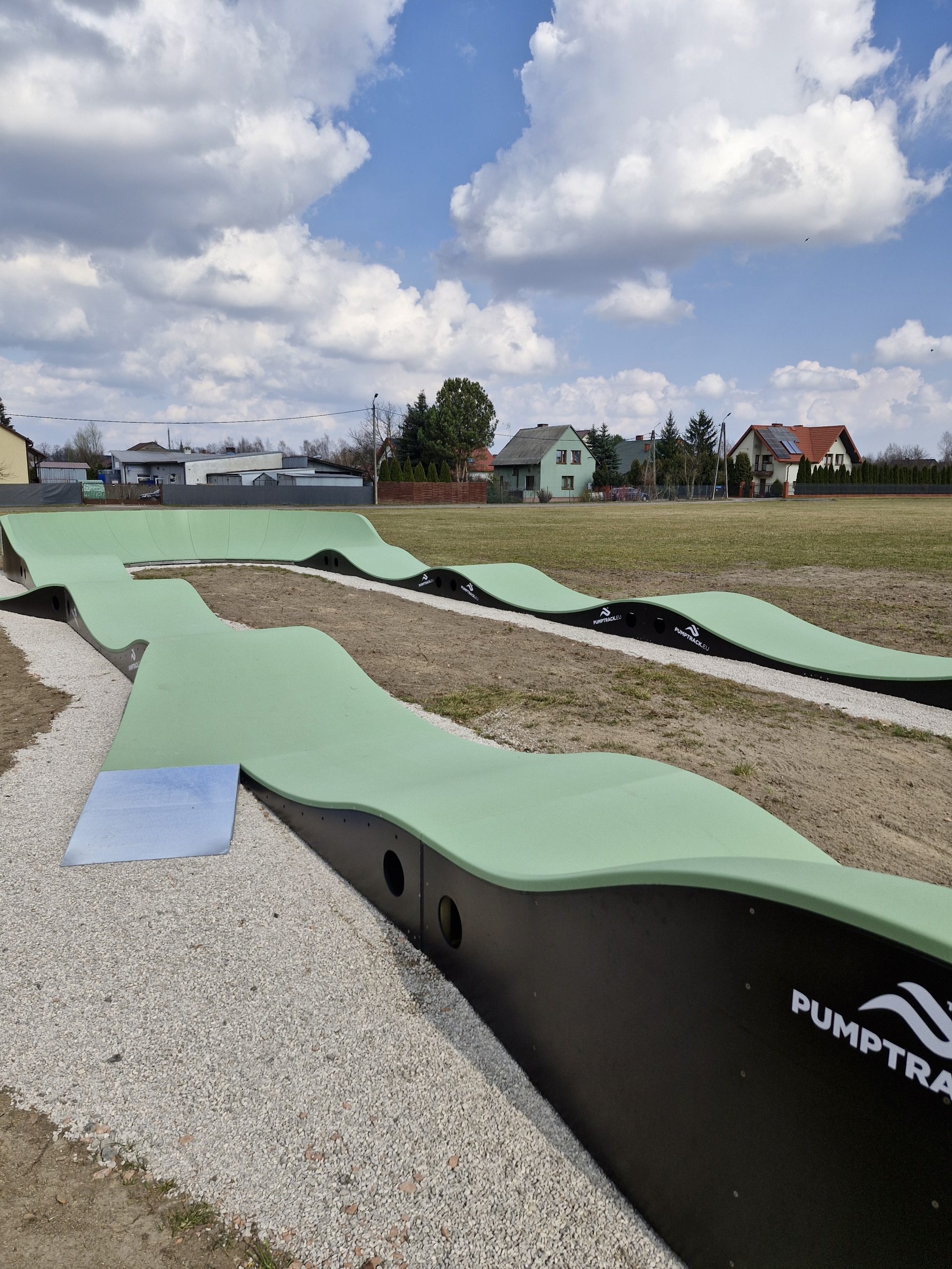 Tor pumptrack o zielonej nawierzchni i czarnych bokach na żwirowej ścieżce. W tle widoczne są domy i drzewa pod niebieskim niebem z chmurami.