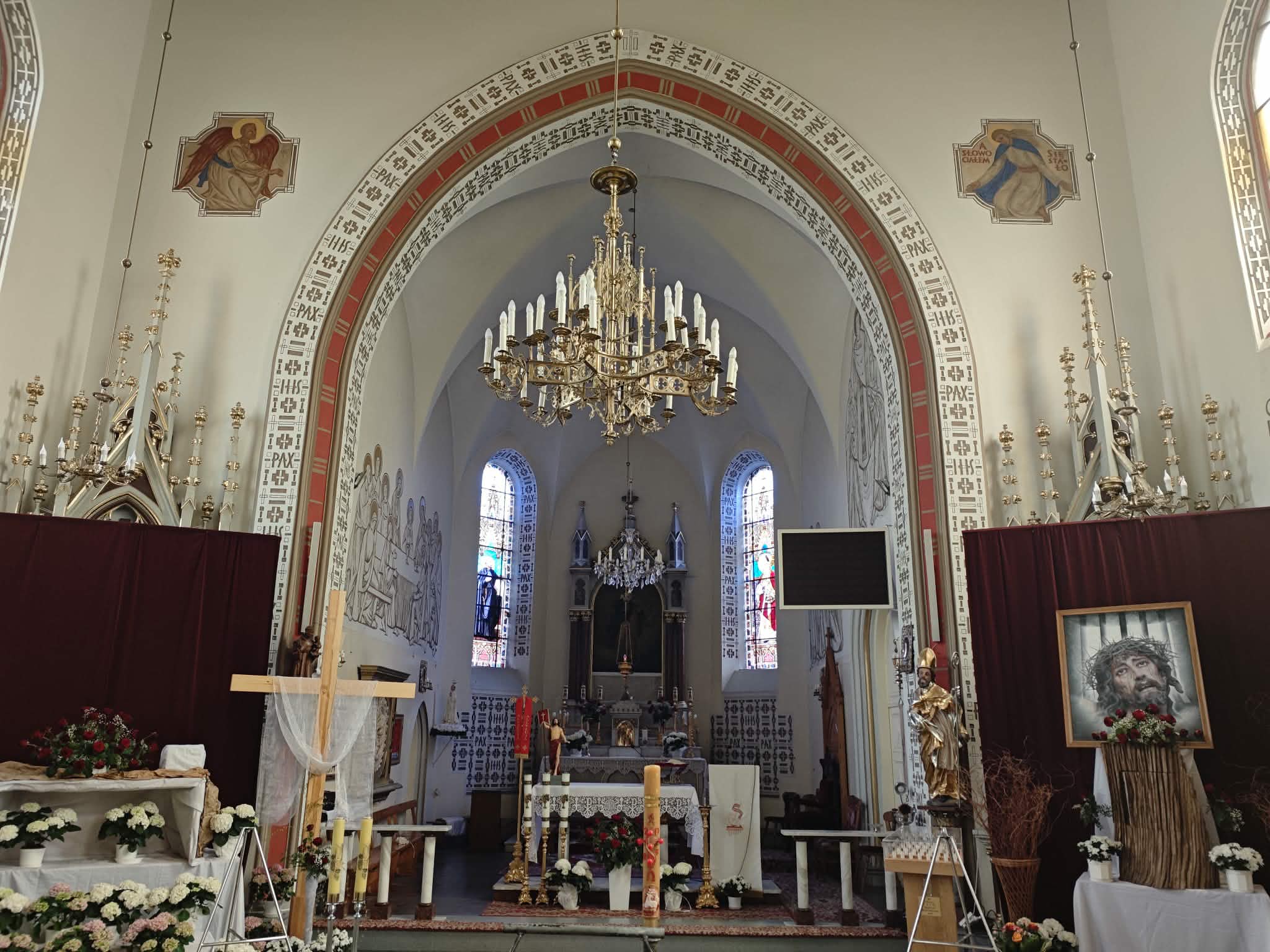 Wnętrze kościoła z ołtarzem, białymi ścianami i złotym żyrandolem. Witraże i religijne obrazy na ścianach. Dekoracje kwiatowe w bieli i czerwieni.