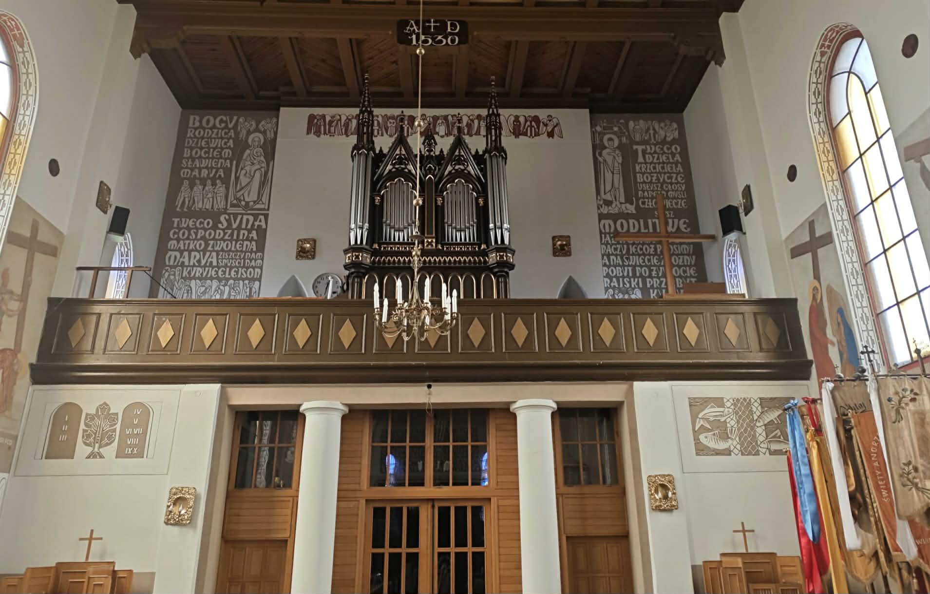 Kościelny chór z organami oraz drewnianym balkonem, zdobiony złotymi wzorami. Tło w neutralnych kolorach z religijnymi napisami. Po bokach widać dekoracje ścienne i flagi.