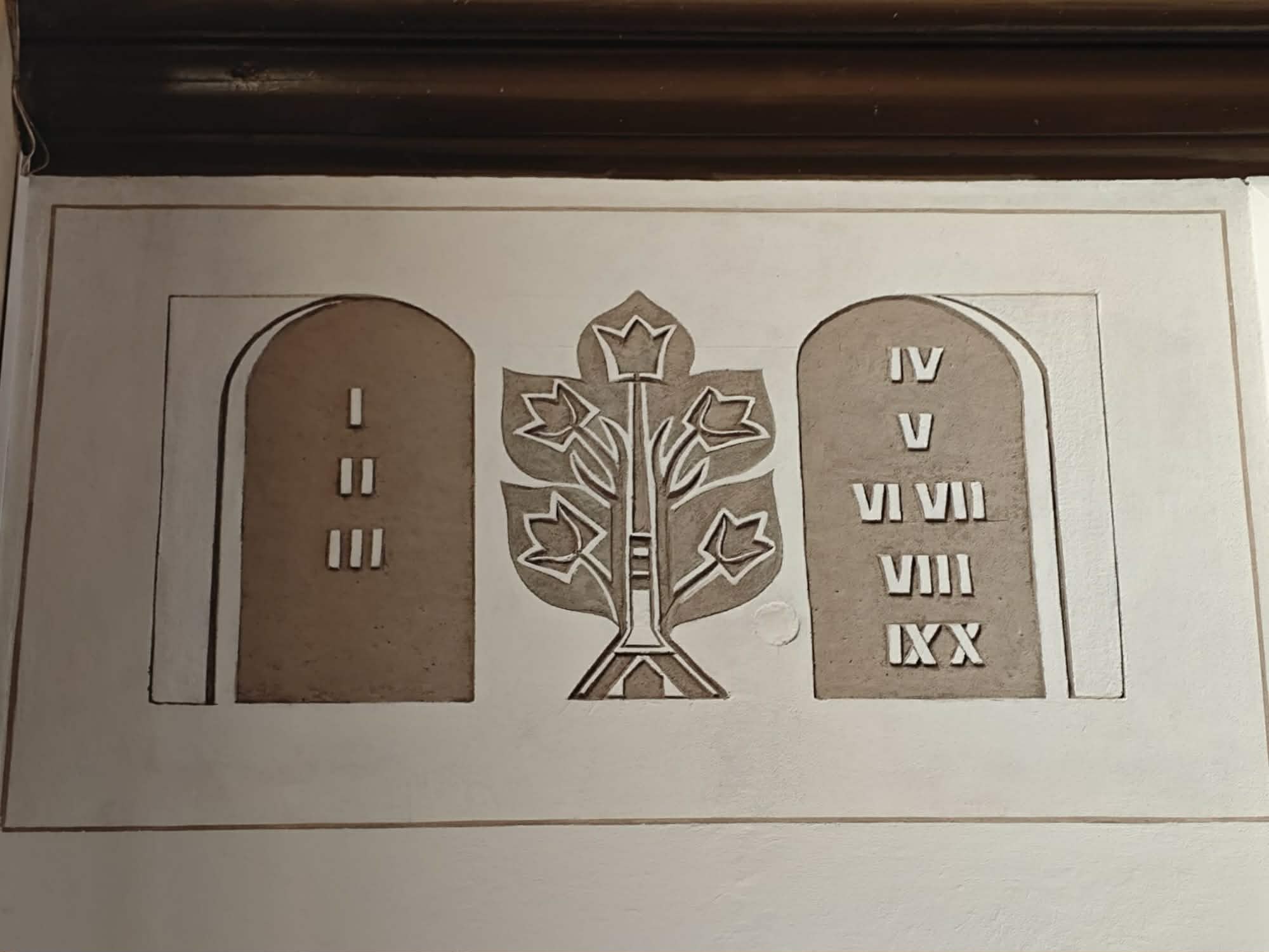 Obraz przedstawia dwie kamienne tablice z numerami rzymskimi i drzewo pośrodku. Kolory to beżowy i biały. W tle widoczna jest biała ściana.