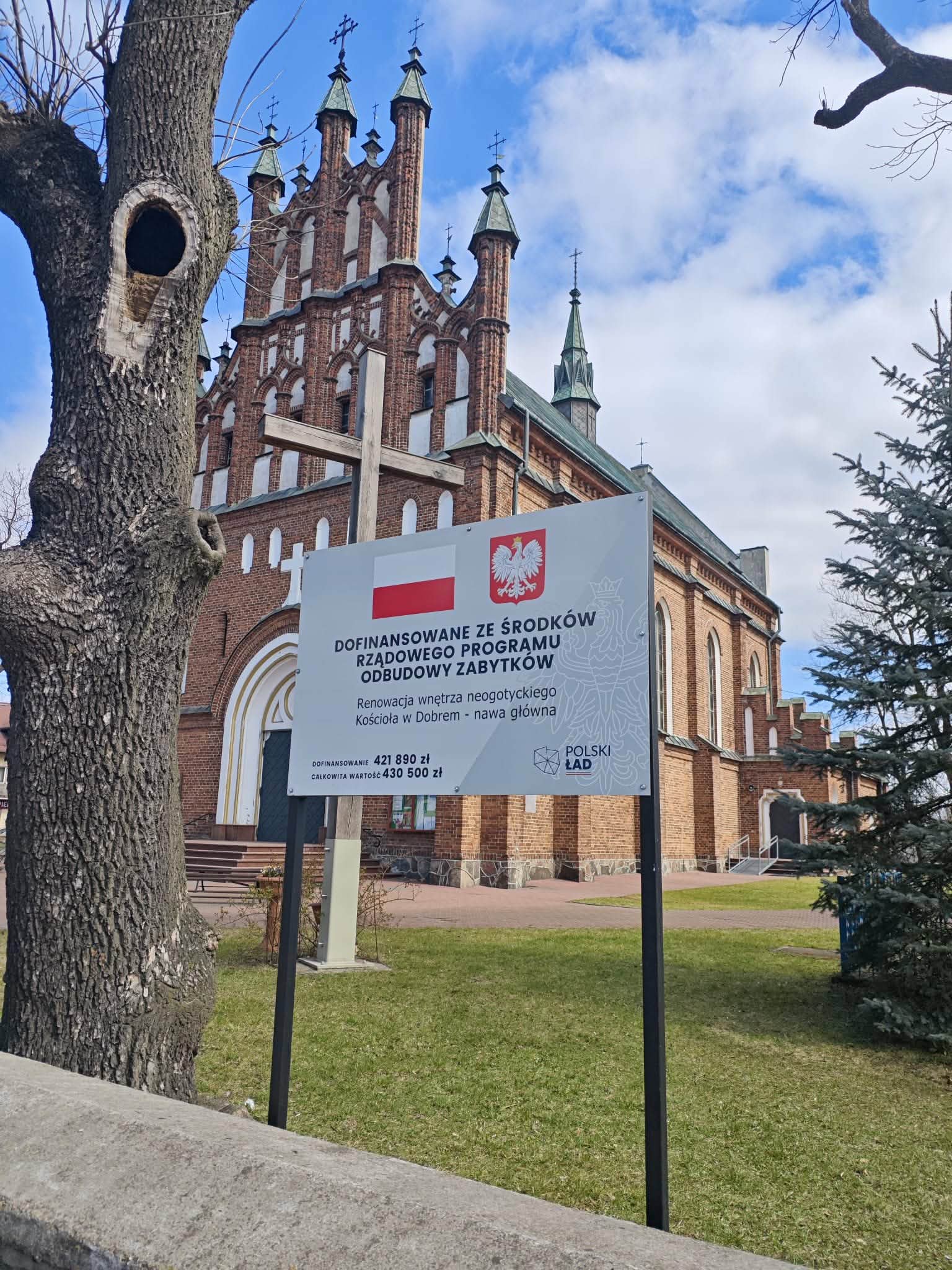 Kościół neogotycki z czerwoną cegłą w tle. Na pierwszym planie drewniany krzyż i tablica informacyjna. Obok drzewo, niebieskie niebo.