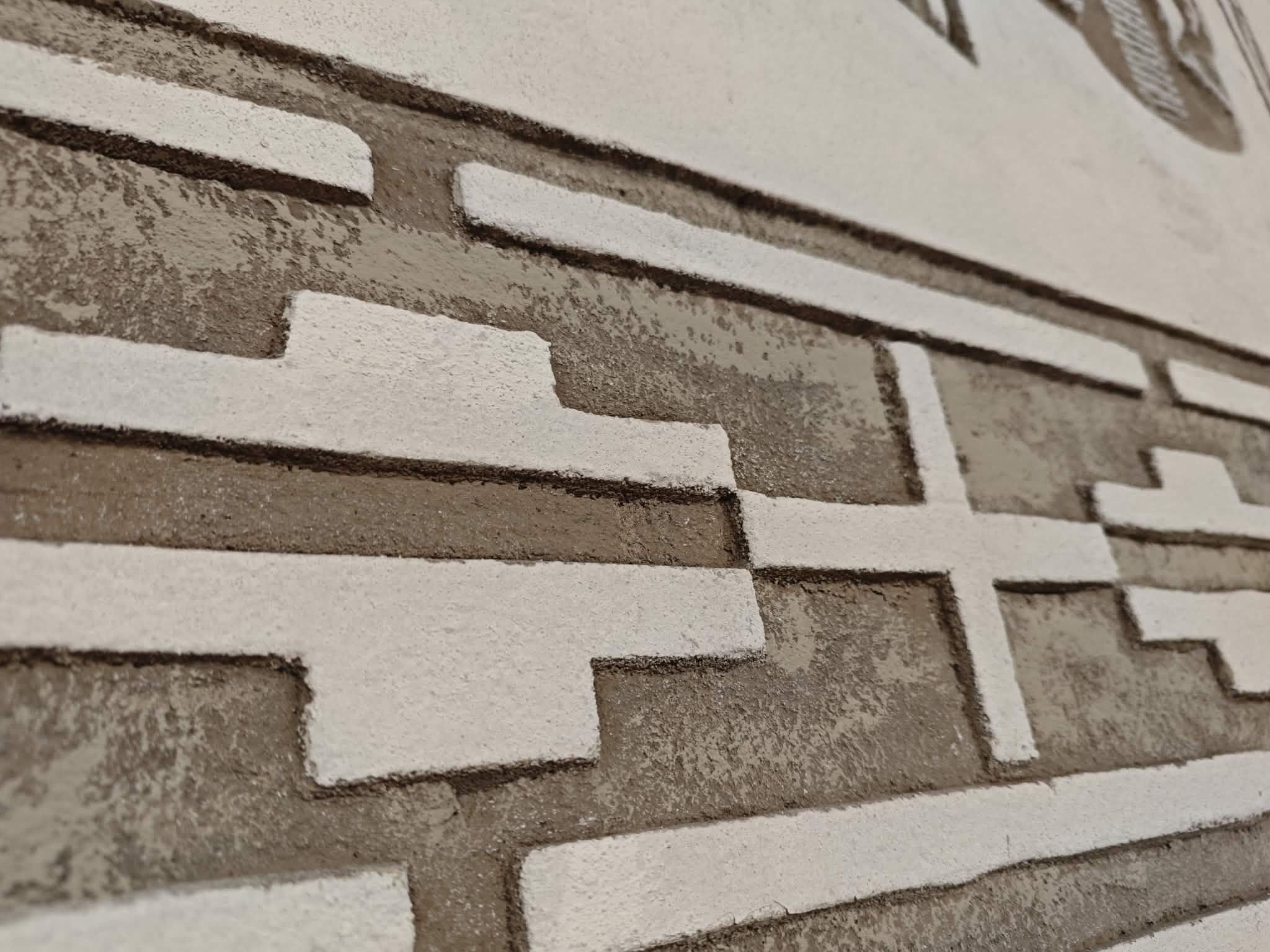 Ściana z geometrycznym wzorem w kolorach beżu i bieli. Struktura jest trójwymiarowa, z wystającymi prostokątami. Tło jasnobeżowe.