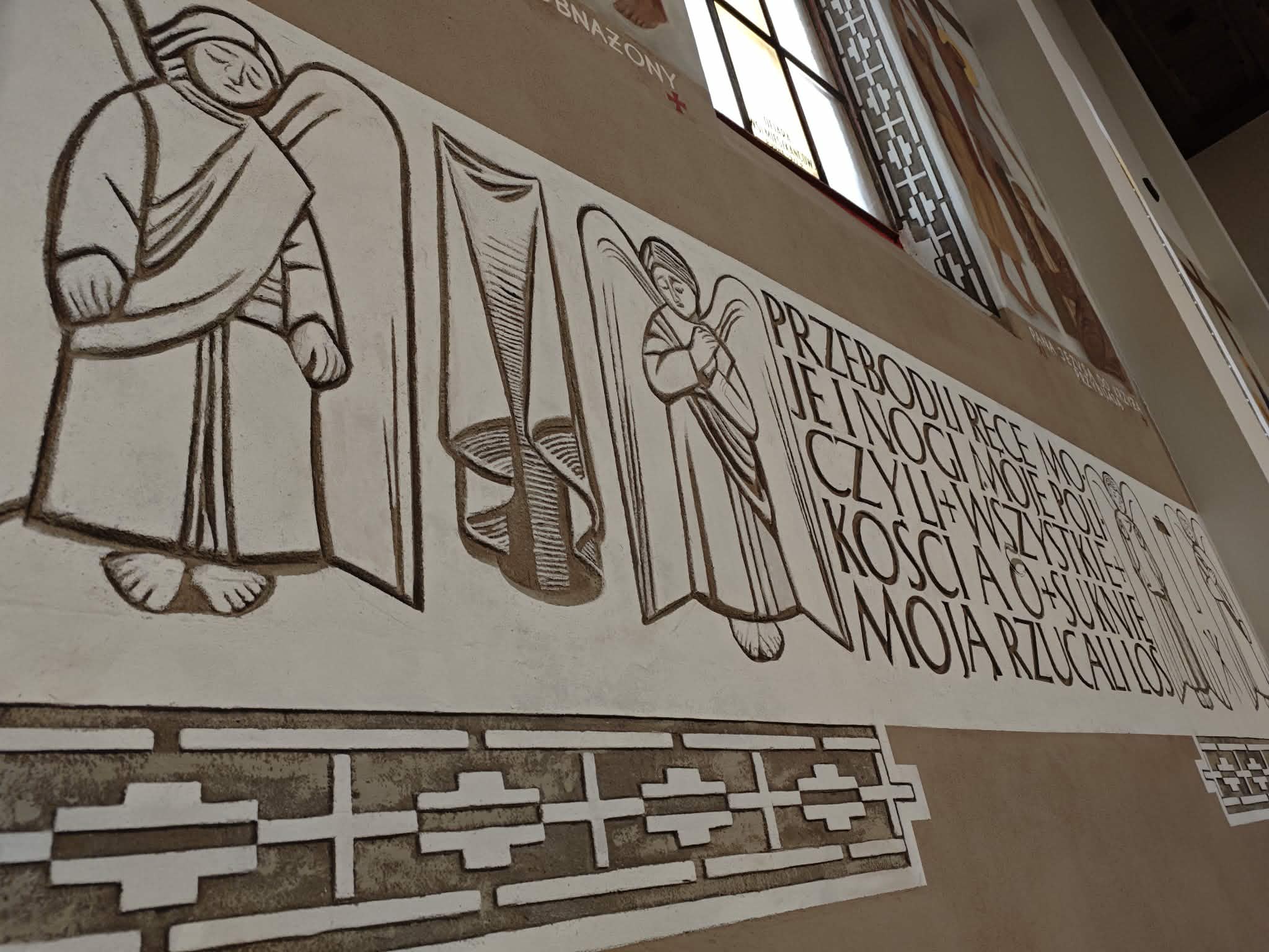 Mur z jasnym freskiem z postaciami aniołów obok napisu w języku polskim. Tło beżowe z ozdobnym białym wzorem. Obok są witraże, wpuszczające światło do wnętrza.