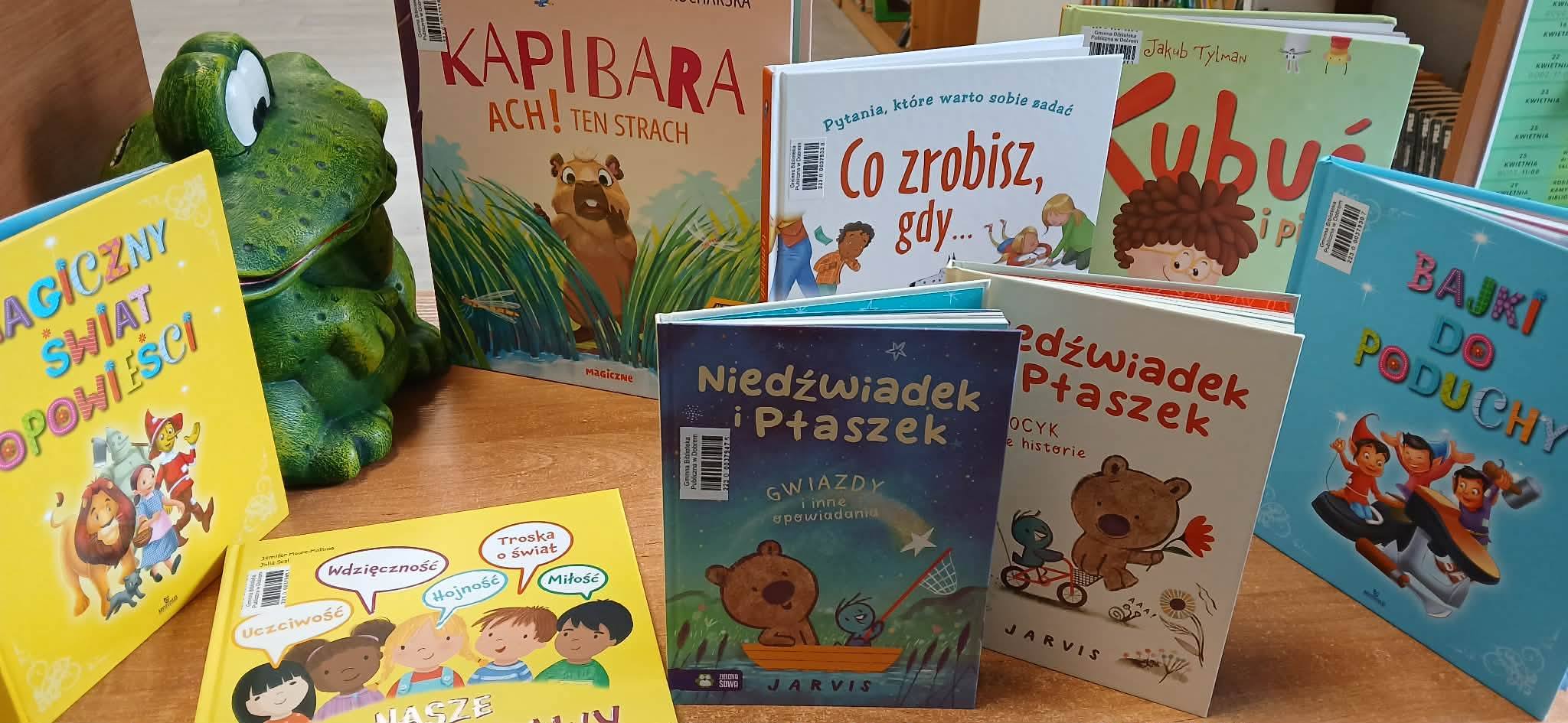 Stos książek dziecięcych w różnych kolorowych okładkach na drewnianym stole, ozdobiony figurką żaby.