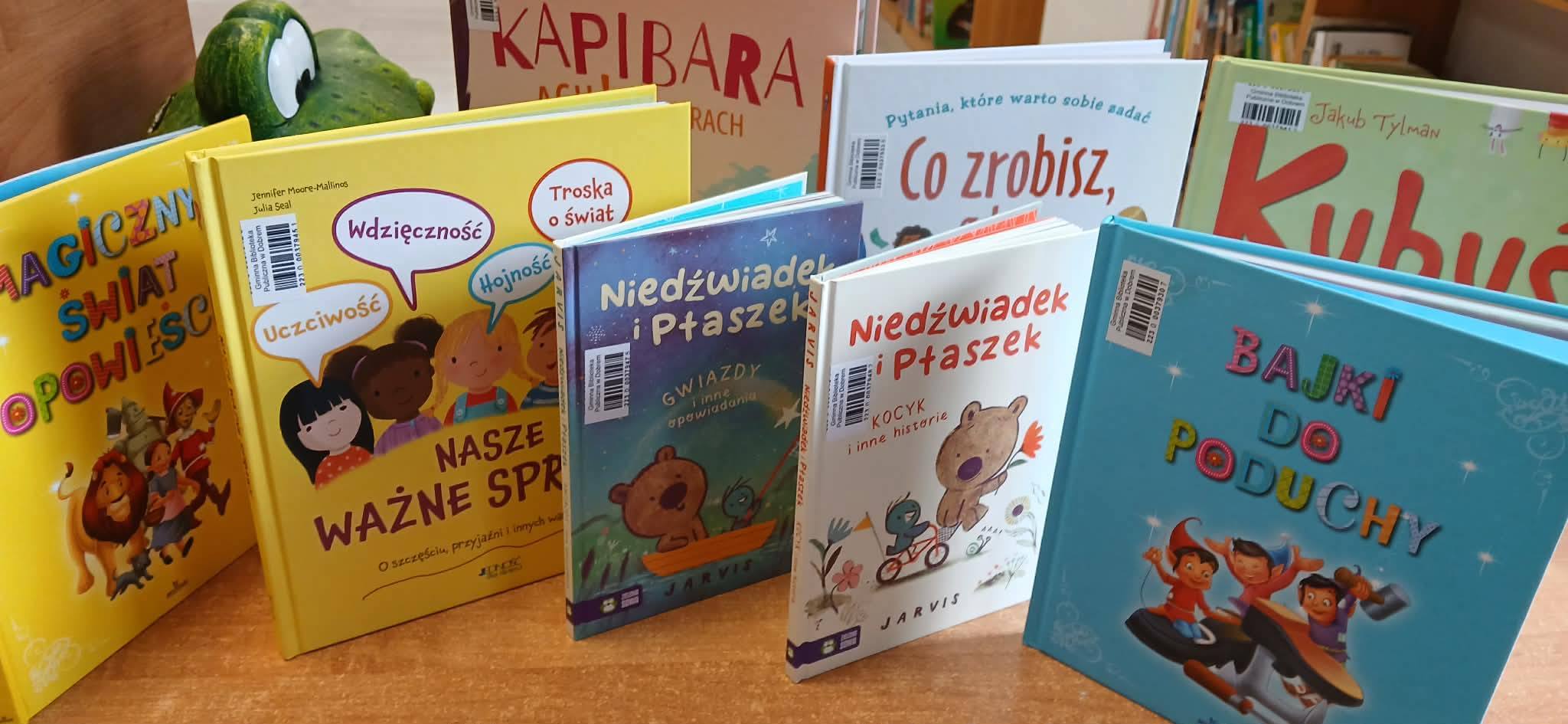 Na zdjęciu widoczne są kolorowe książki dla dzieci na drewnianej półce. Okładki w jasnych barwach, z dużymi, kolorowymi napisami i ilustracjami. W tle znajduje się zielona dekoracja w kształcie lisa.