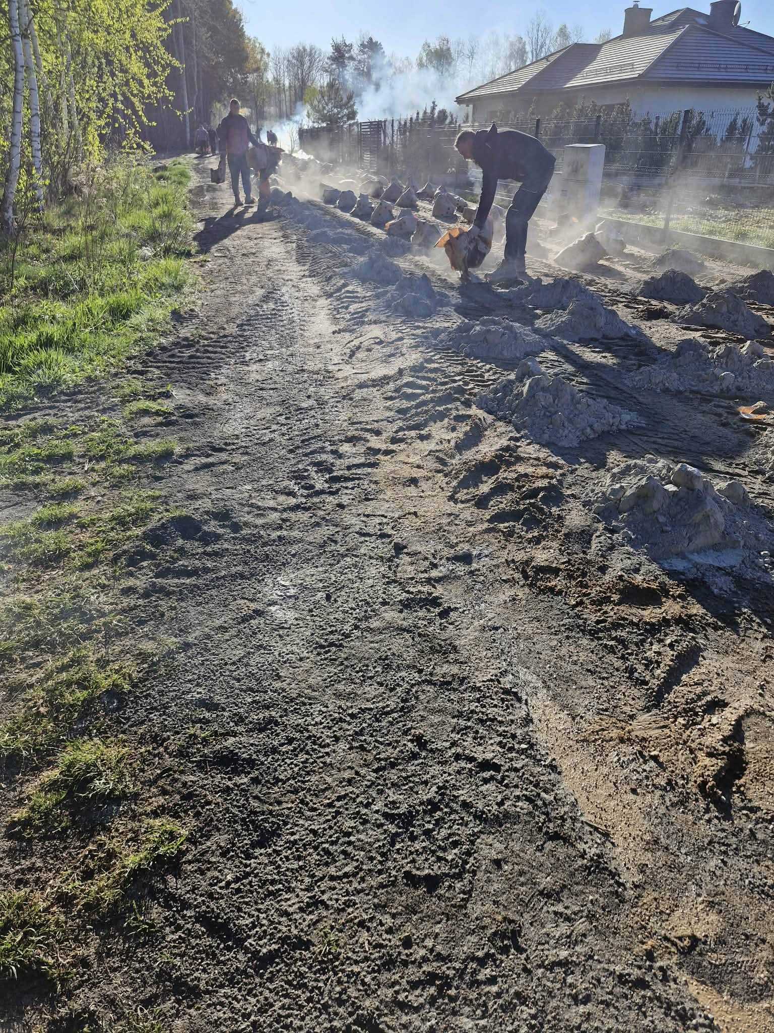 Pracownicy naprawiają gruntową drogę, podnosząc kłęby kurzu. Obok zielone drzewa i dom z białym płotem. Jasne, słoneczne popołudnie.