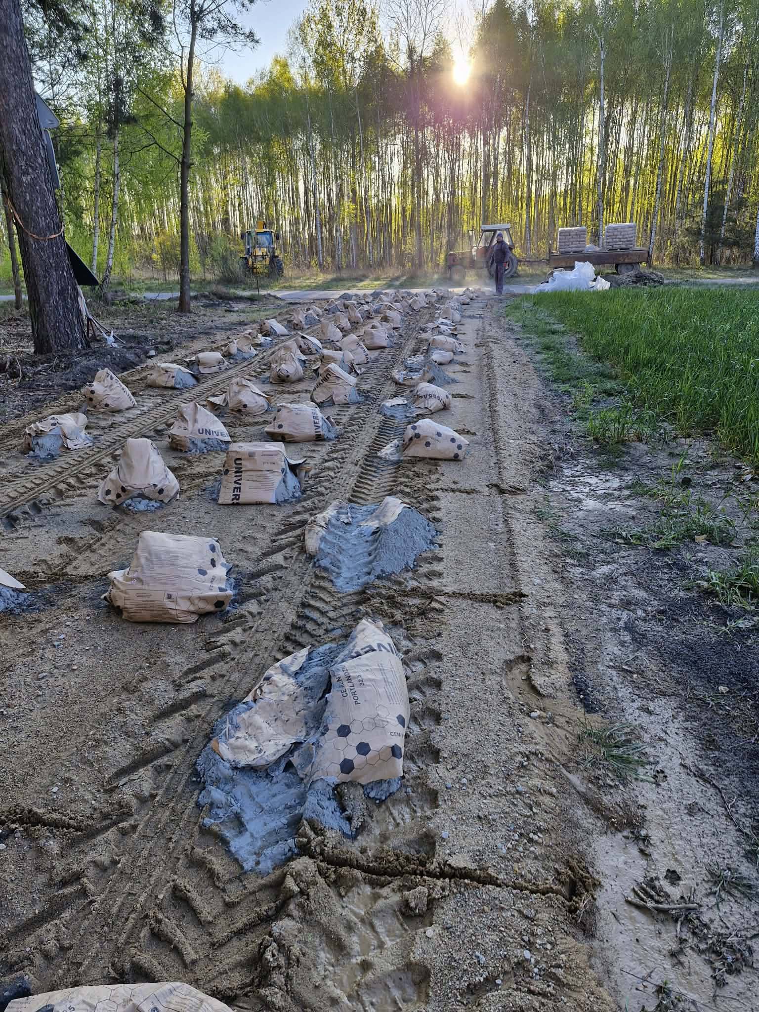 Na zdjęciu widoczne są worki z cementem na piaszczystej drodze. Tło stanowi las w odcieniach zieleni oświetlony zachodzącym słońcem. Po prawej stronie stoi traktor z przyczepą załadowaną materiałami.