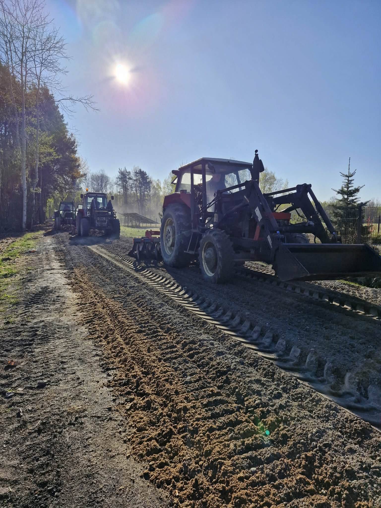 Traktory jadą drogą gruntową obok lasu, słońce świeci jasno na niebie. Ziemia jest wzruszona i pokryta śladami opon. W tle drzewa i czyste niebo.