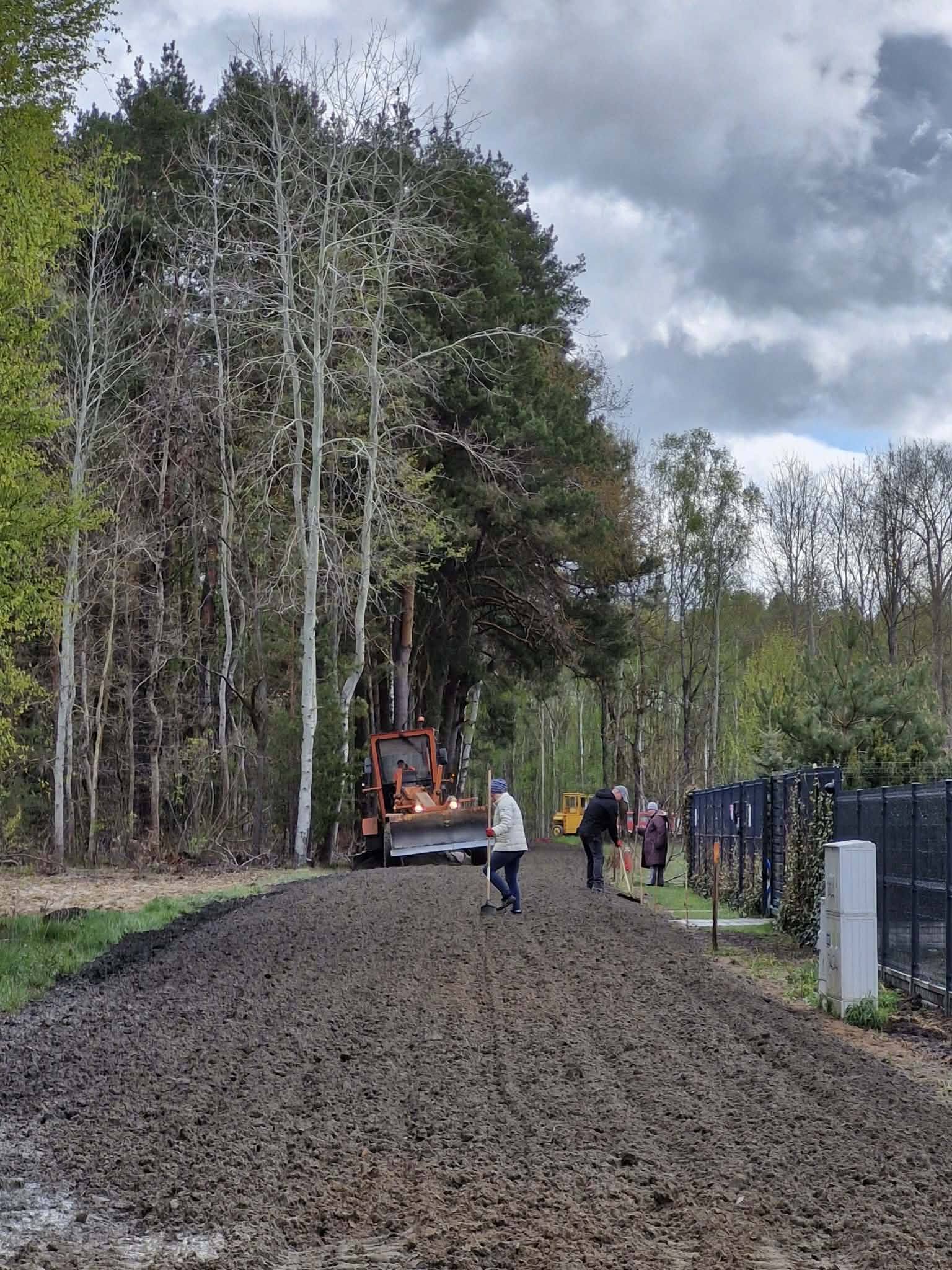 Na zdjęciu widoczny jest traktor na drodze otoczonej drzewami, pracujący nad utwardzaniem powierzchni. Tło to zieleń lasu i szare niebo z chmurami. Kilku ludzi z narzędziami pochyla się nad ziemią.