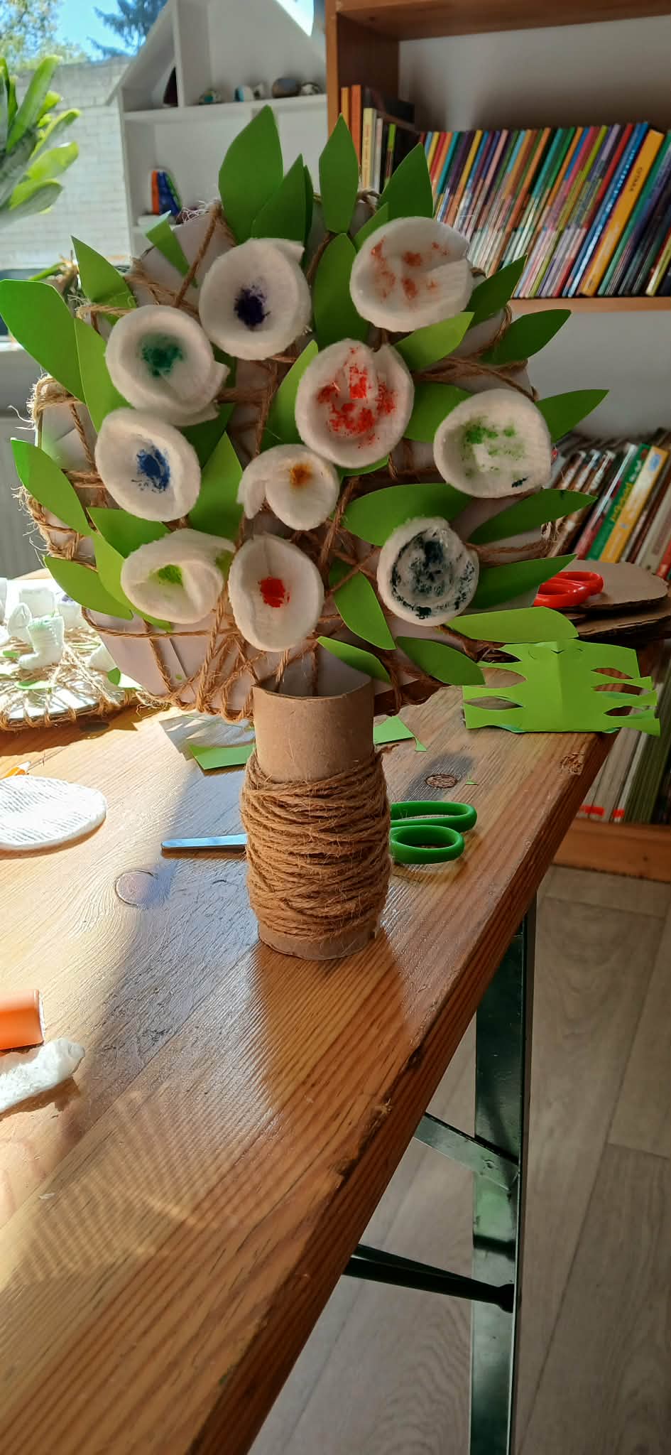 Na drewnianym stole stoi ręcznie wykonane drzewko z papieru i sznurka. Liście są zielone, a kwiaty mają kolorowe środki. W tle półki z książkami.