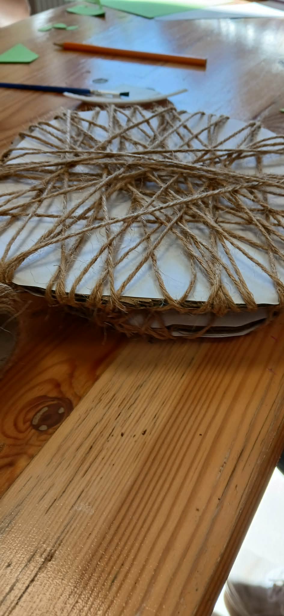 Na drewnianym stole leży biały przedmiot opleciony sznurkiem jutowym. W tle widoczne są zielone i pomarańczowe kartki. Obraz jest jasny i pełen naturalnych kolorów.