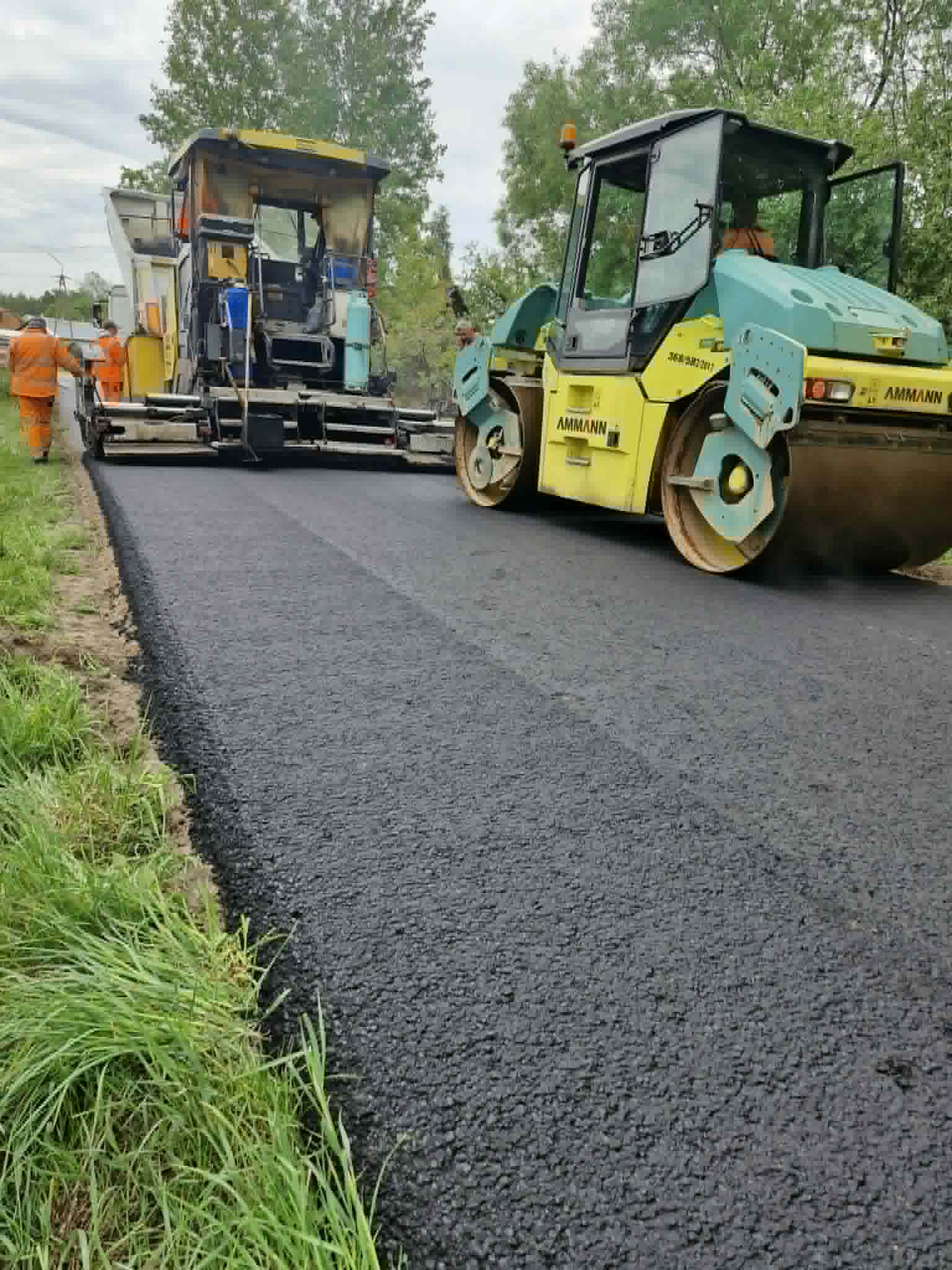 Droga asfaltowana przez maszynę, po lewej dwaj pracownicy w pomarańczowych kombinezonach. Obok zielono-żółty walec. W tle zielone drzewa.