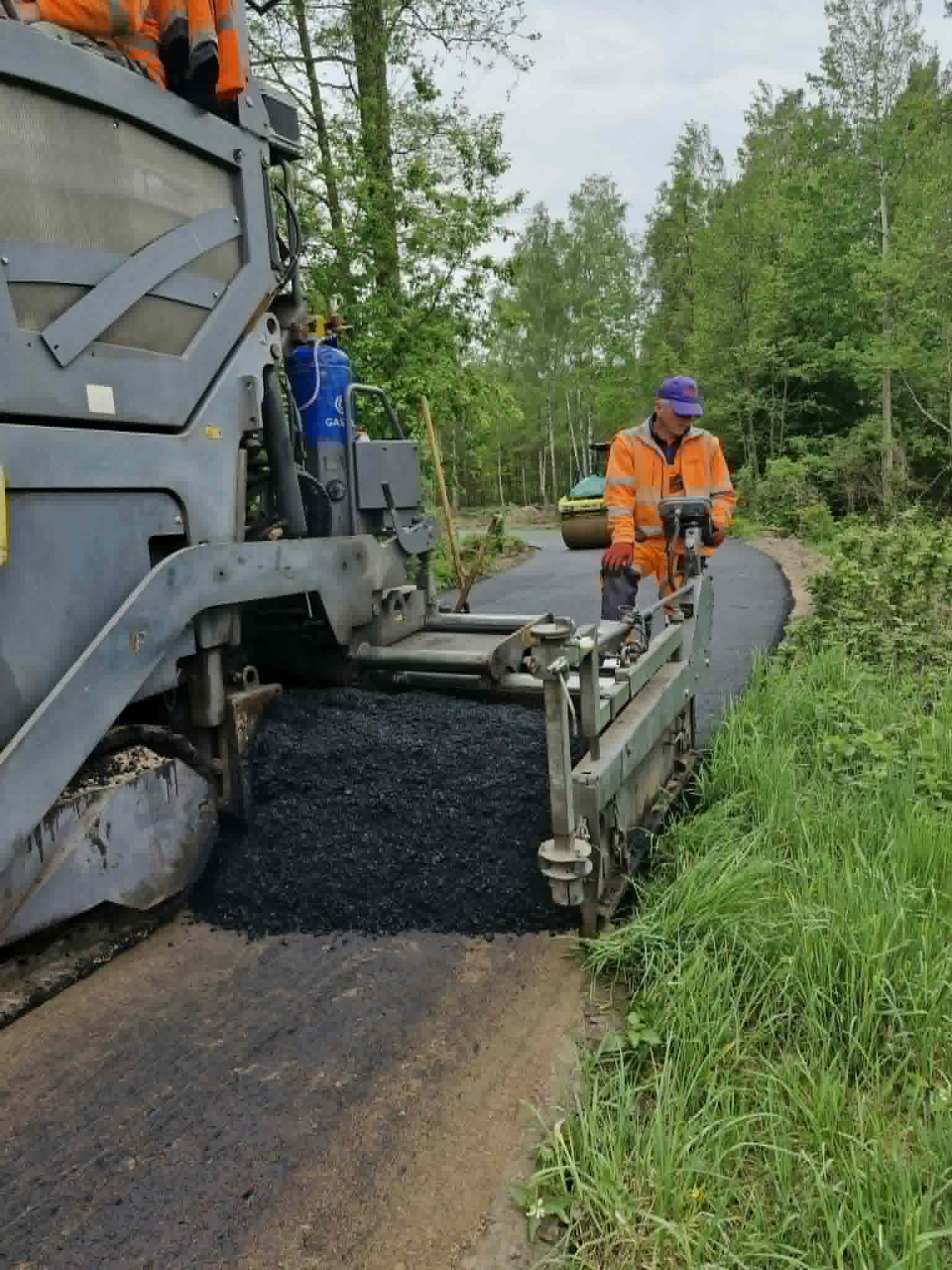Robotnik w pomarańczowym stroju obsługuje asfaltowanie drogi. Szara maszyna kładzie świeży asfalt. W tle zielone drzewa i trawa.