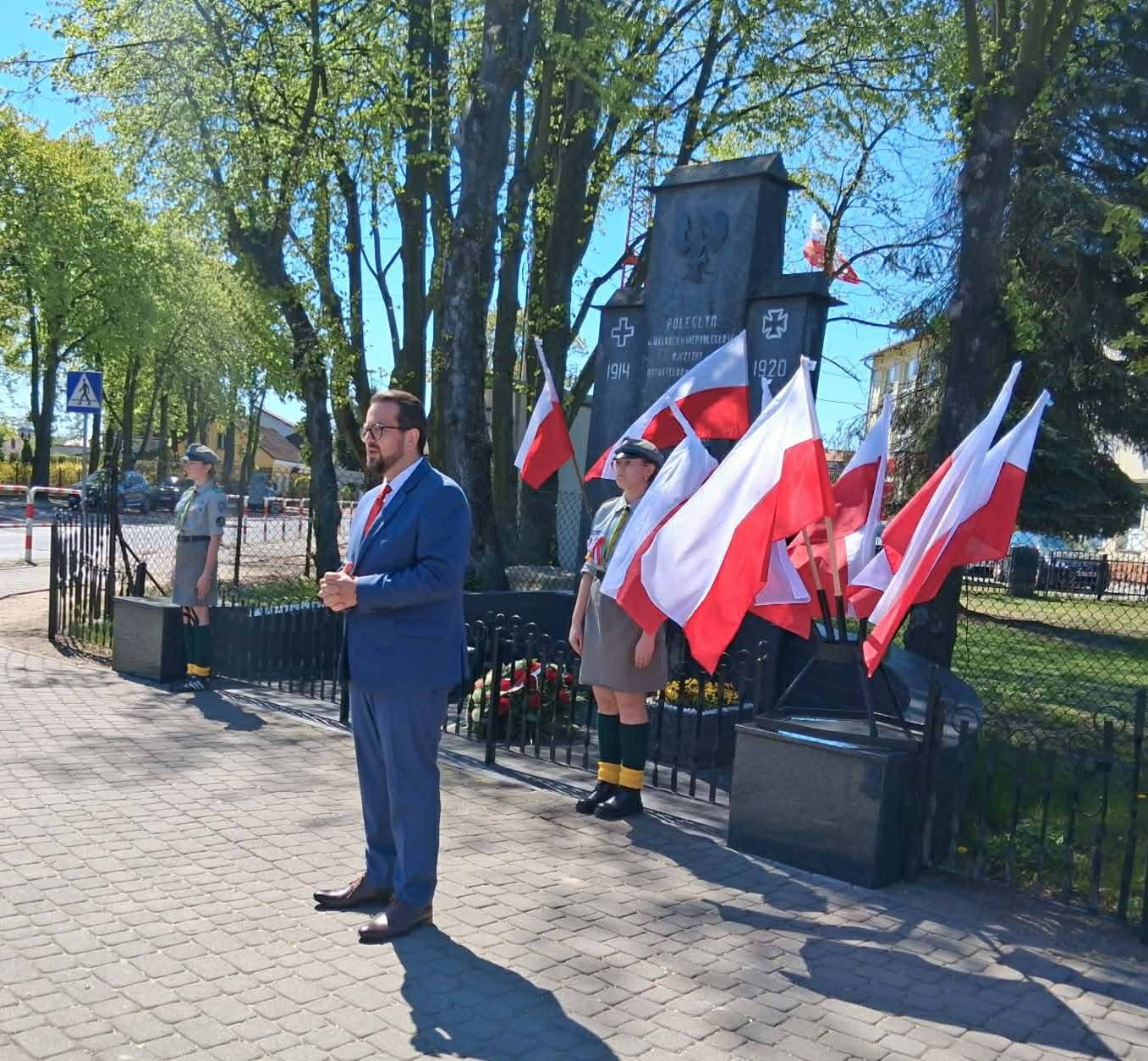 Mężczyzna w niebieskim garniturze stoi przed pomnikiem otoczonym polskimi flagami. Dwie osoby w mundurach harcerskich stoją obok. Tło tworzą drzewa i jasne, słoneczne niebo.