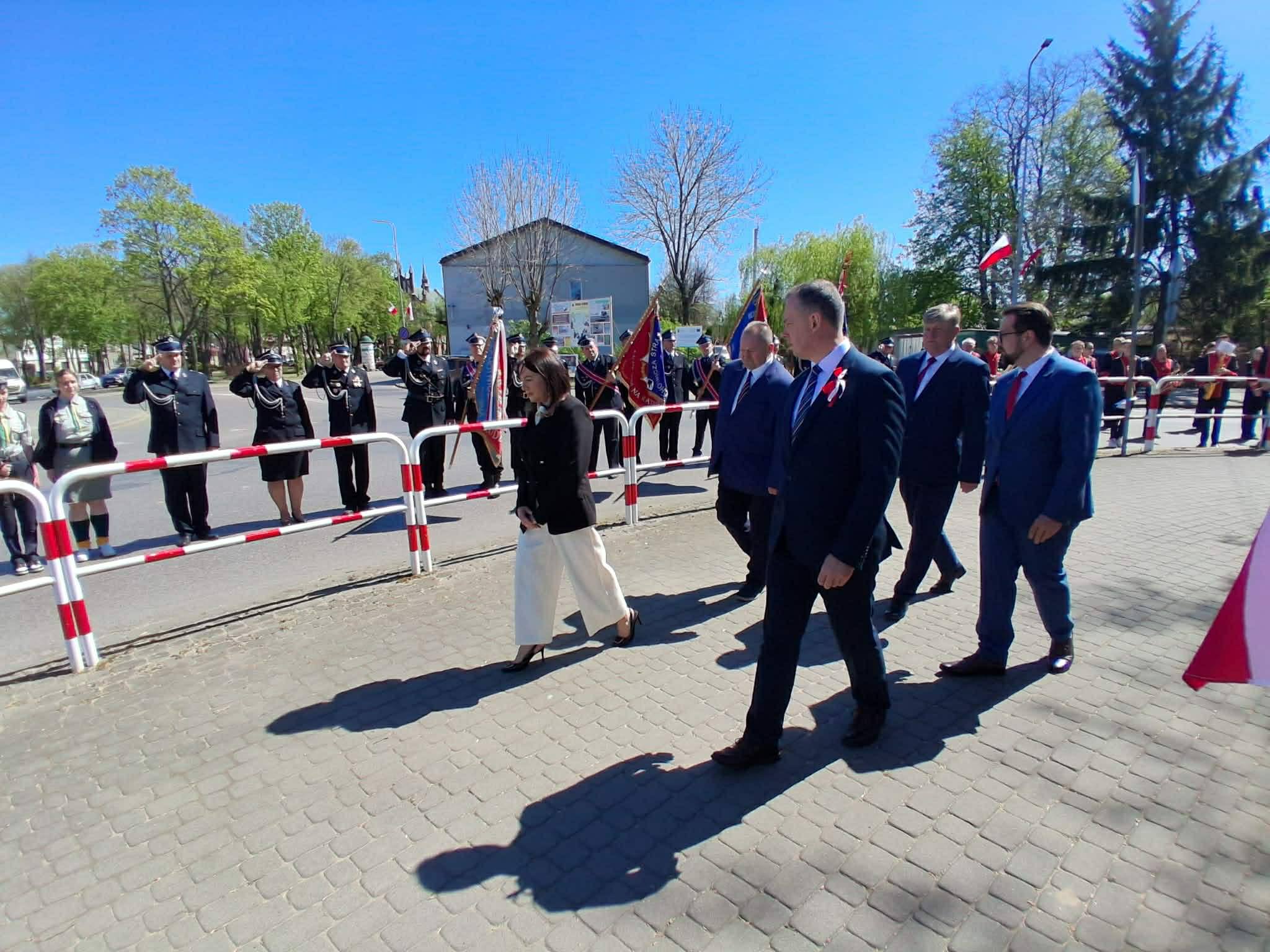 Grupa osób w eleganckich strojach idzie chodnikiem. W tle znajdują się strażacy w mundurach i flagi. Jest słoneczny dzień, a niebo jest czyste.