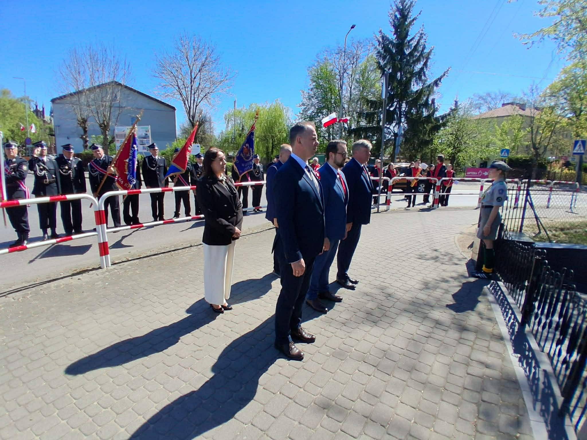 Na zdjęciu widoczni są ludzie w eleganckich strojach w otoczeniu pocztów sztandarowych. Tło stanowią zielone drzewa i niebieskie niebo. Wydarzenie odbywa się na zewnątrz, na brukowanej powierzchni.