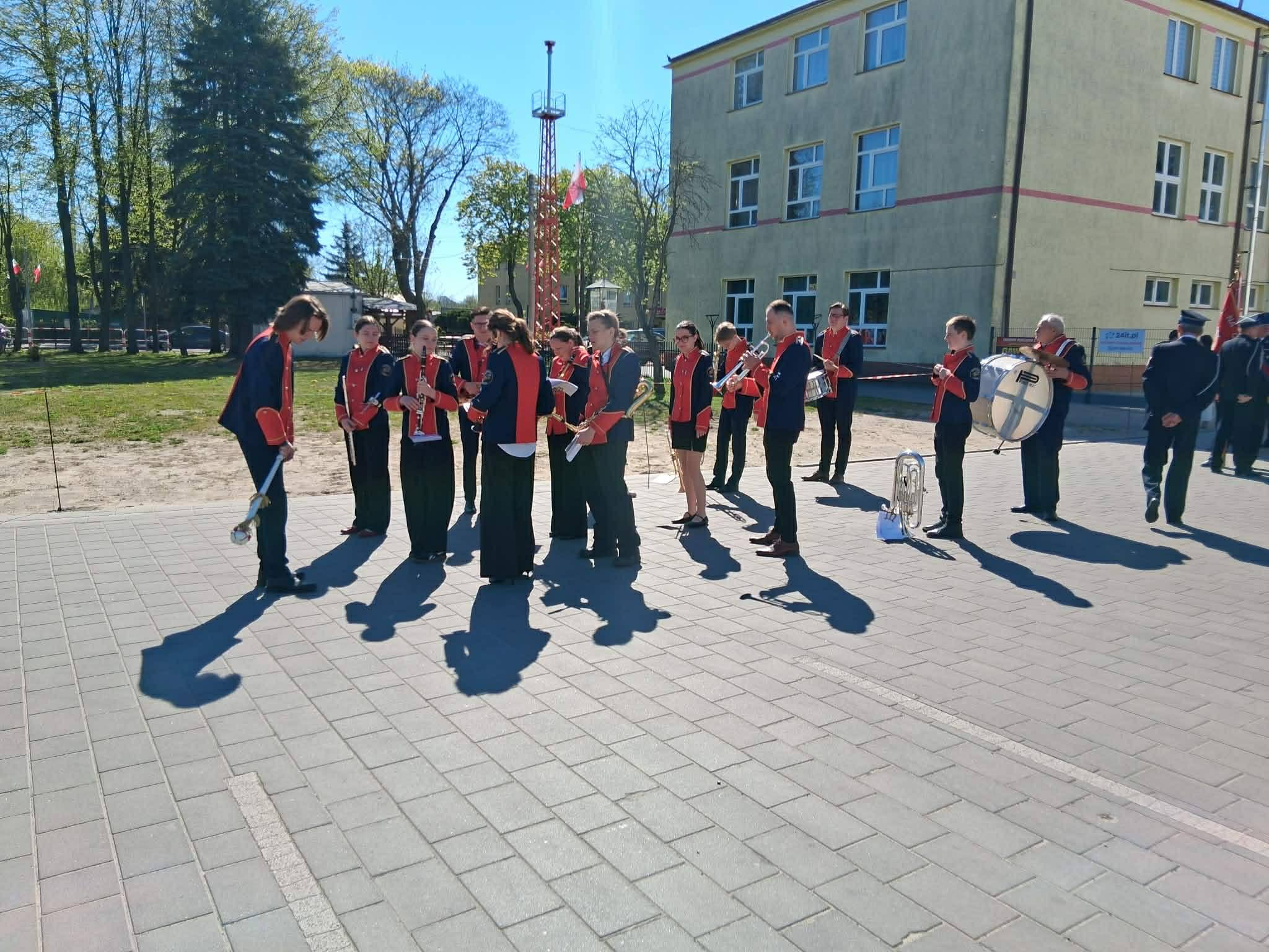 Orkiestra w czerwonych strojach gra na placu. Tło stanowi żółty budynek i drzewa pod jasnym niebem. Cień muzyków kładzie się na chodniku.