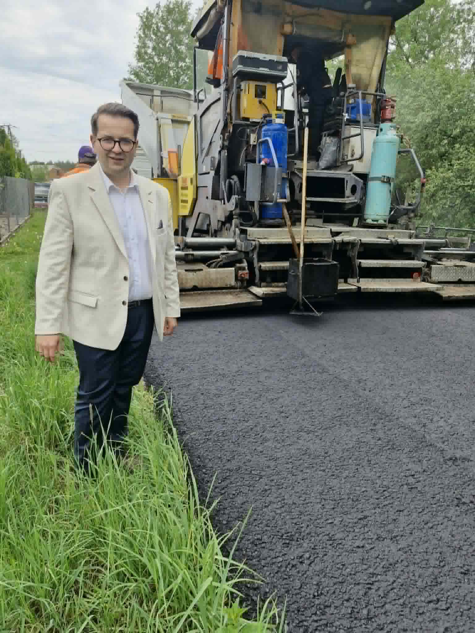 Na zdjęciu mężczyzna w jasnej marynarce stoi obok świeżo wyłożonego asfaltu. Obok niego znajduje się maszyna do asfaltowania. Otoczenie to zielona trawa i drzewa w tle.
