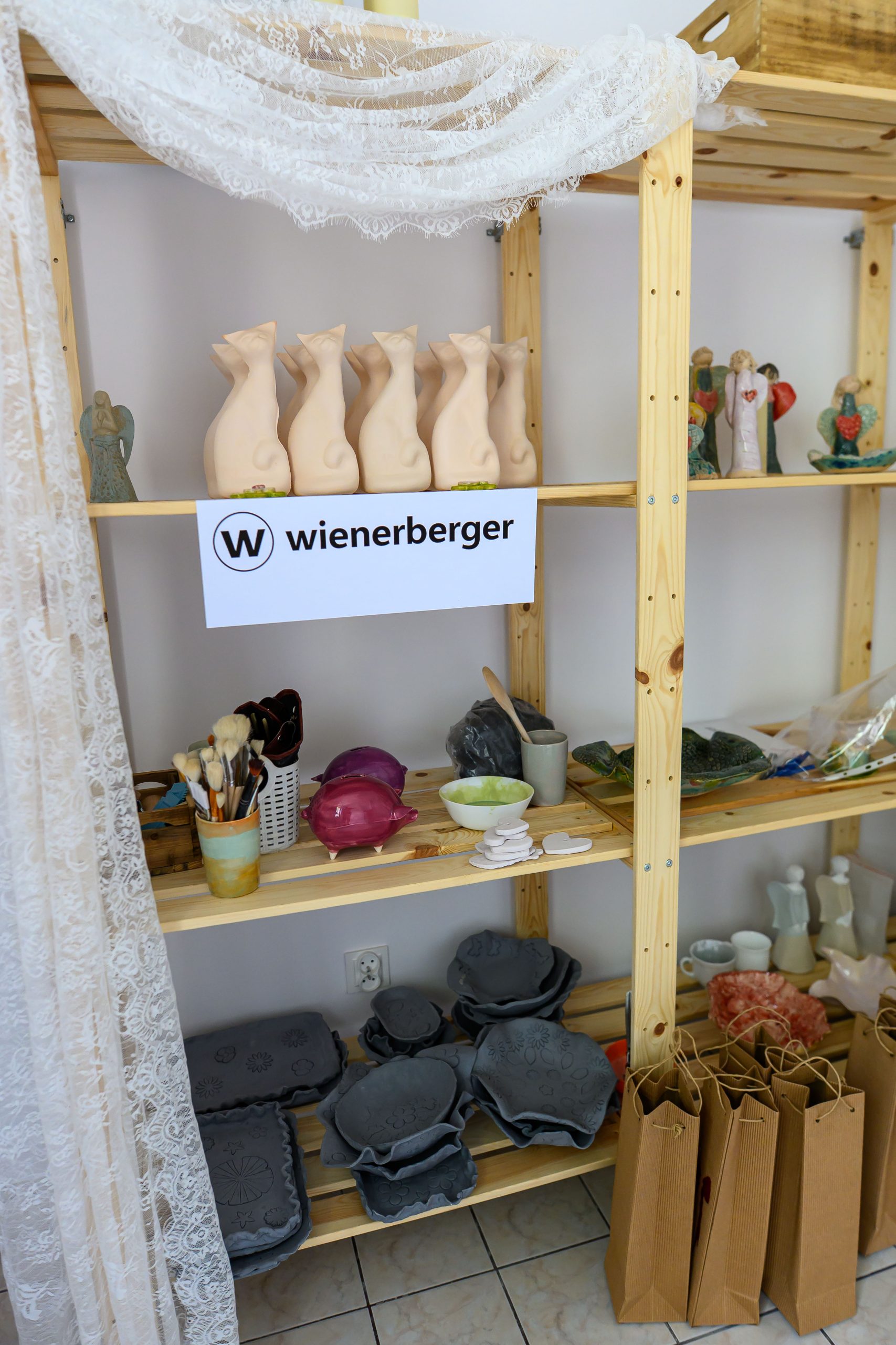 Drewniane półki z glinianymi figurkami i malarskimi przyborami na tle białej ściany. Dominują beże, biel i szarość. Na środkowej półce logo Wienerberger.