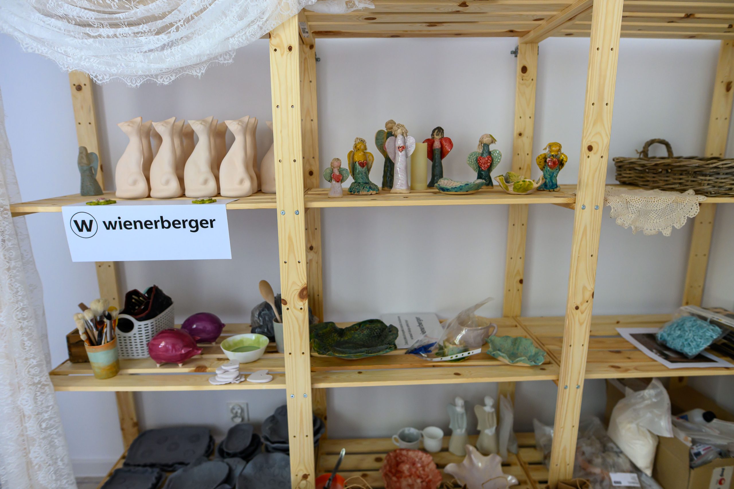 Drewniany regał z ceramicznymi figurkami i materiałami rzemieślniczymi. Kolory są stonowane, dominują beże i zielenie. Obok znajduje się kosz i białe zasłony.