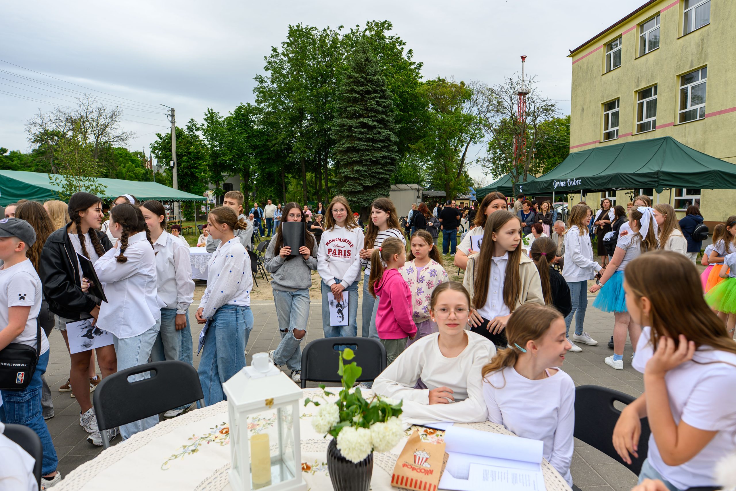 Grupa młodzieży w jasnych ubraniach stoi na zewnątrz przed budynkiem. W tle zielone namioty i drzewa, pogodny dzień. Na pierwszym planie stół z białym obrusem, świecznikiem i popcornem.