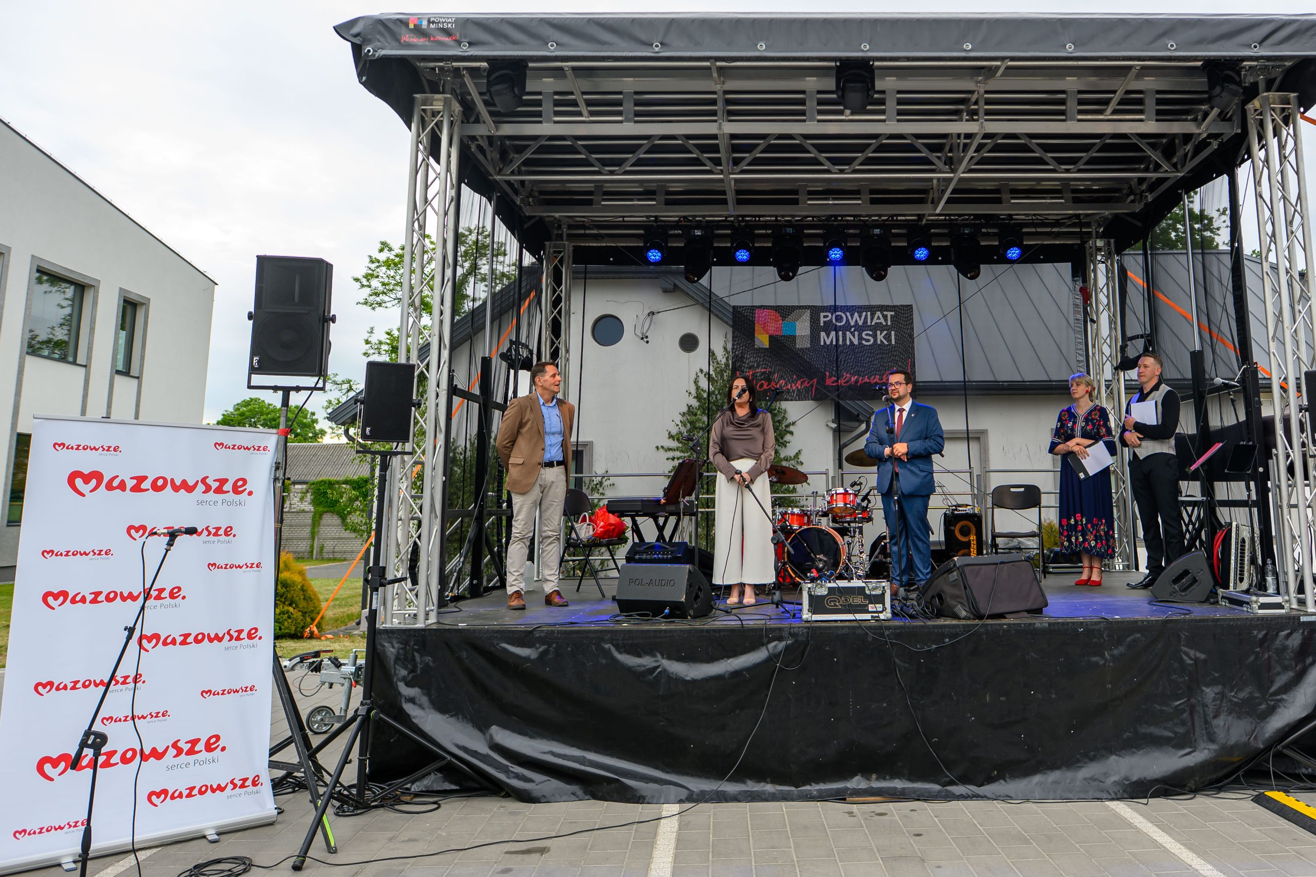 Scena z kilkoma osobami, stoisko z logo Mazowsza po lewej. Tło z szarym budynkiem, czarny dach nad sceną. Kolory głównie stonowane.