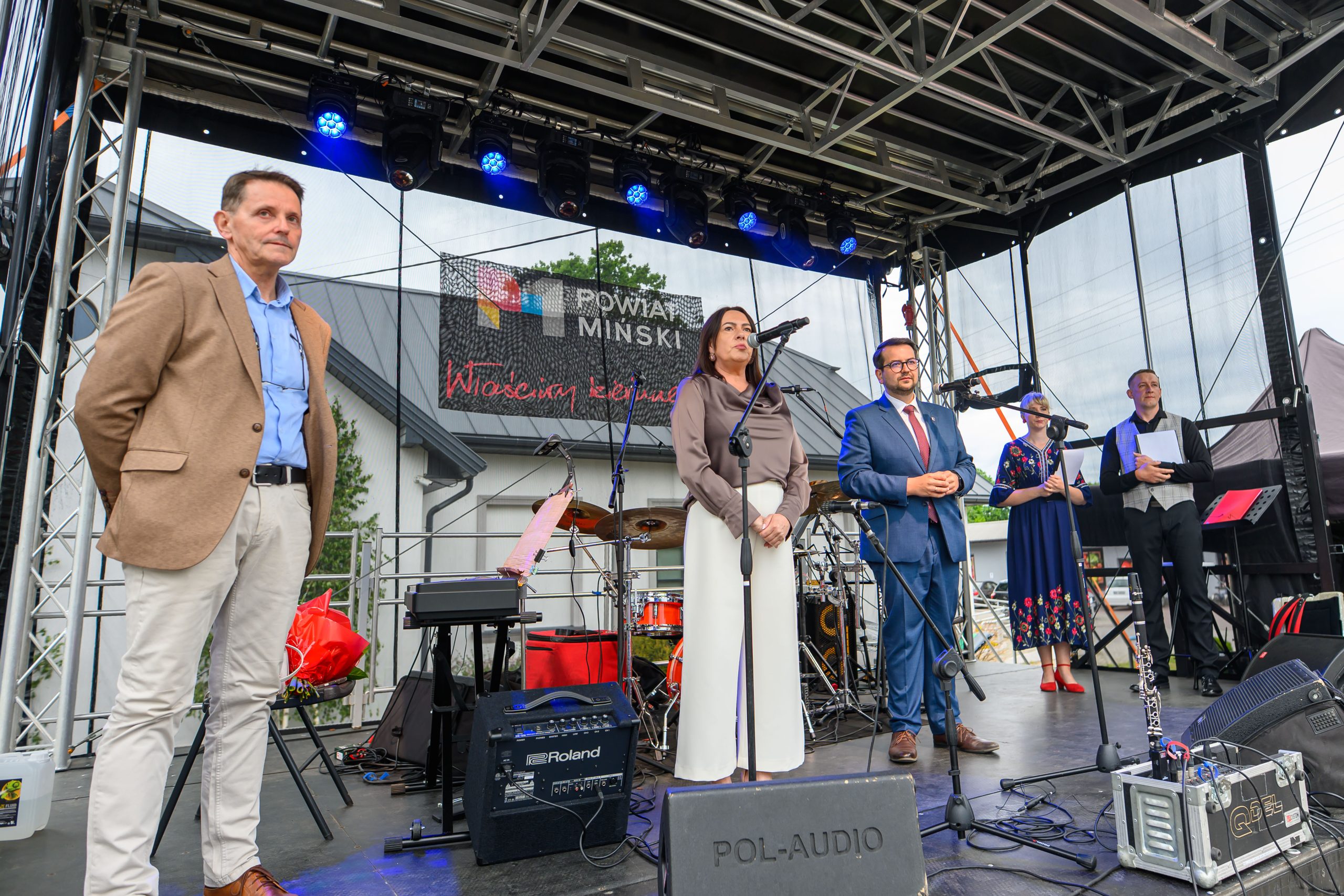 Na scenie stoi pięć osób przemawiających do mikrofonu. Tło stanowi baner z logiem i napisem Powiat Miński. Scena jest zadaszona, wokół sprzęt audio i instrumenty.