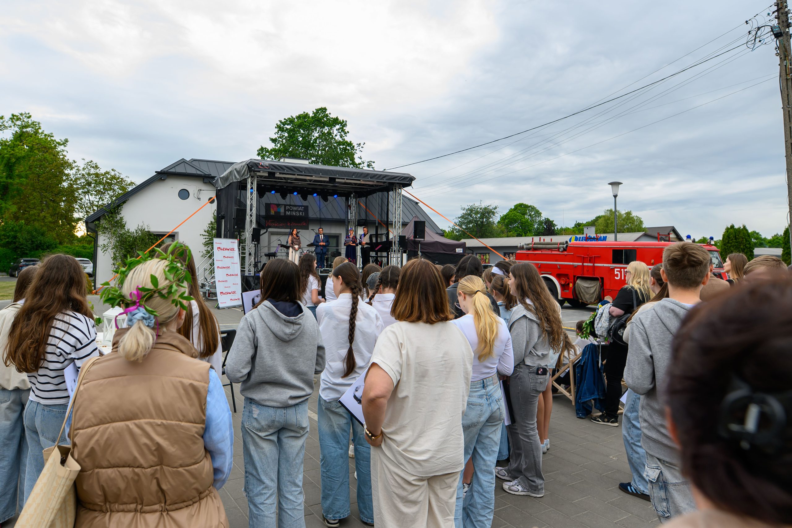 Na zdjęciu jest plenerowy koncert z zespołem na scenie. Widownia składa się z młodych ludzi w jasnych ubraniach. W tle widać budynek, drzewa i czerwony wóz strażacki.