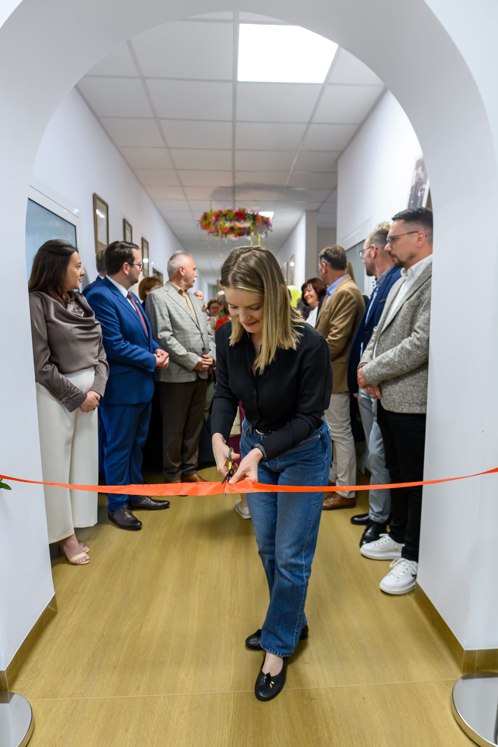 Chociaż nie mogę rozpoznawać osób, ten obraz pokazuje kobietę przecinającą czerwoną wstęgę w korytarzu. Otacza ją grupa ludzi w formalnych ubraniach. Tło jest jasne, z białymi ścianami.
