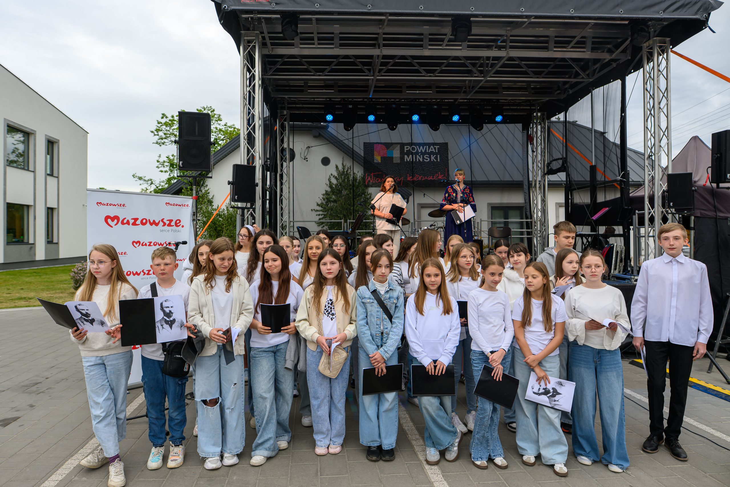 Na zdjęciu jest grupa dzieci ubranych na biało i błękitno przed sceną z dwiema osobami. Tło to nowoczesny budynek i zielona trawa. Nad sceną napis Powiat Miński.
