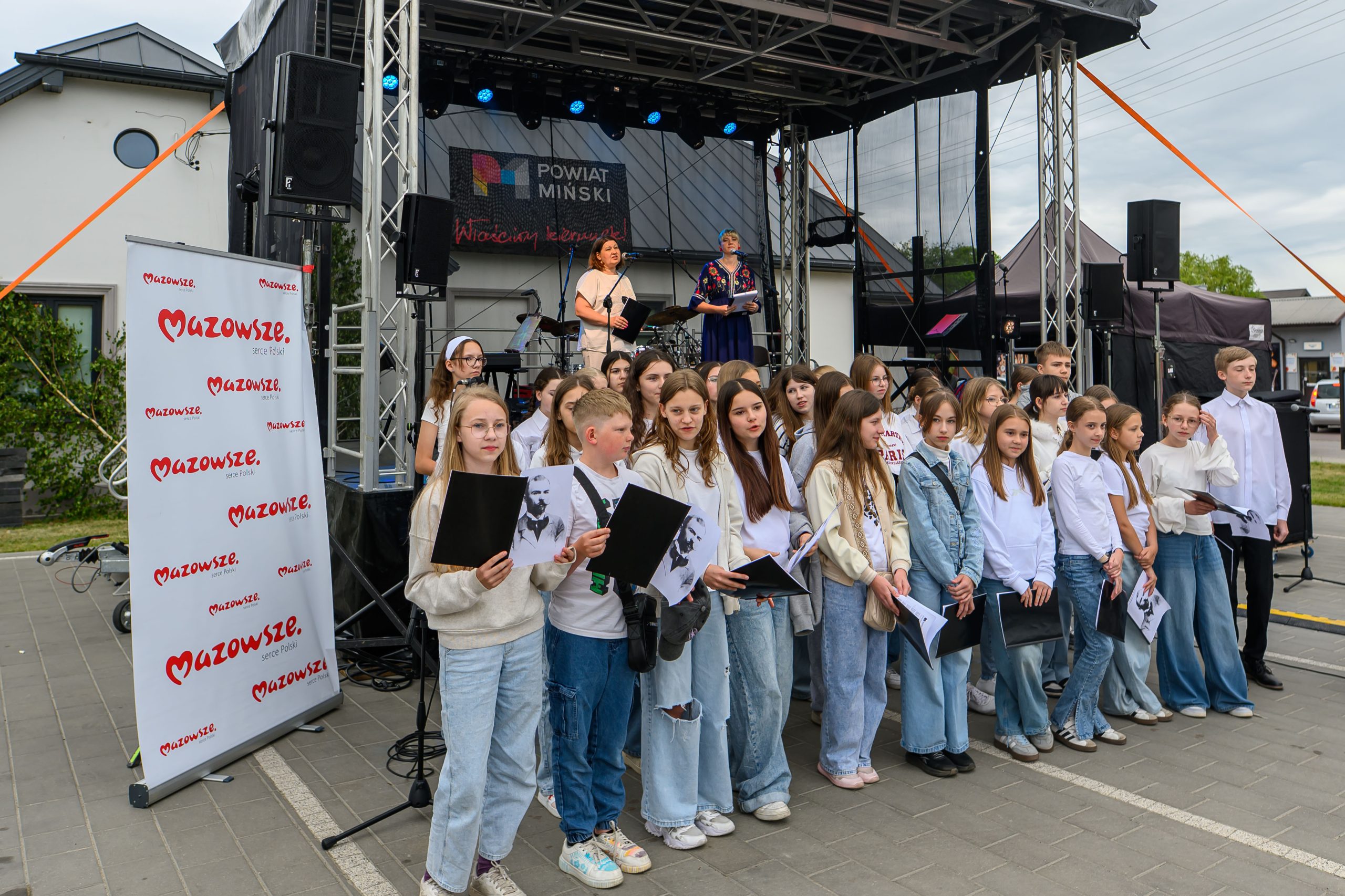 Grupa dzieci w białych i niebieskich strojach stoi przed sceną. W tle plakat Mazowsze. Sceneria plenerowa, scena z występem.