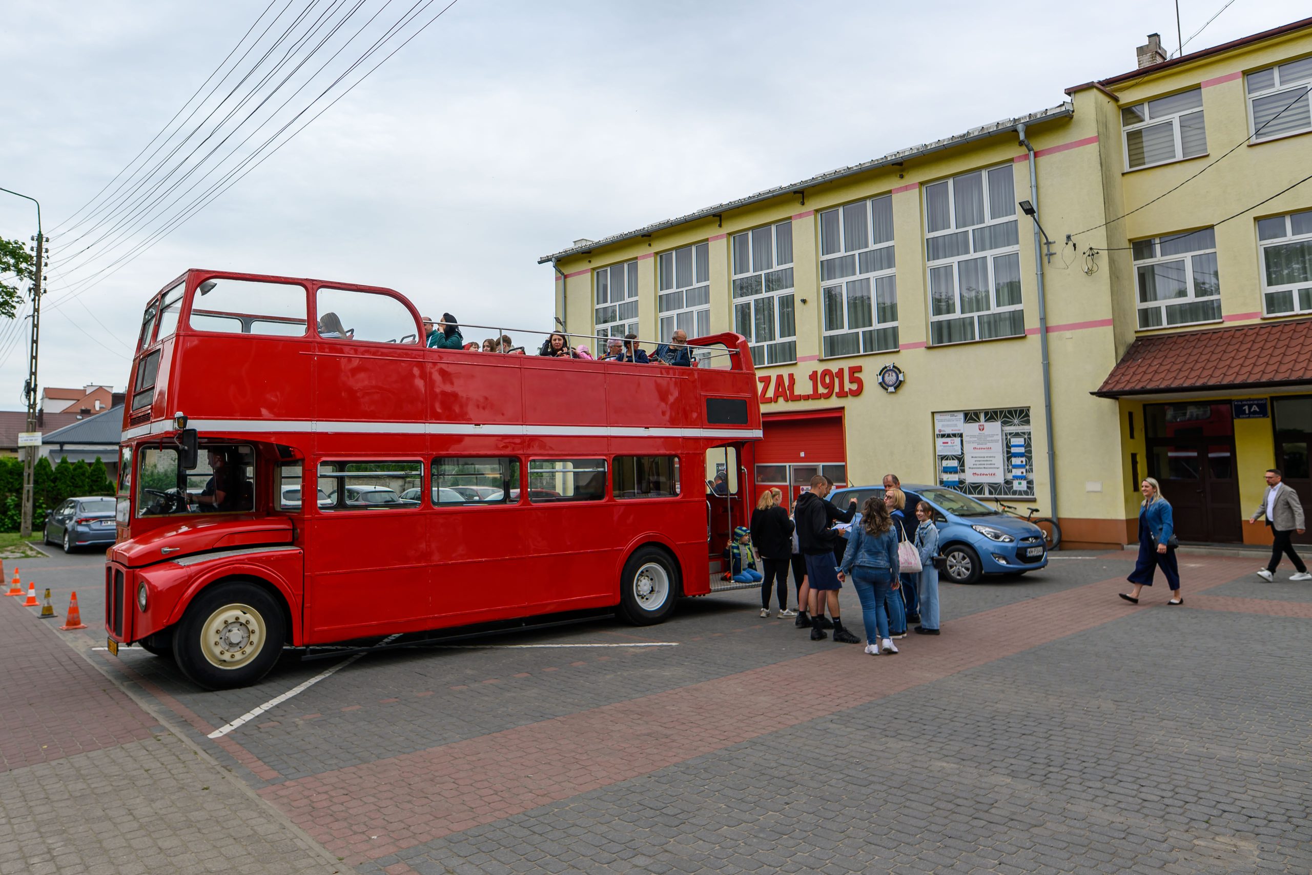 Czerwony piętrowy autobus stoi przed żółtym budynkiem z napisem OCHOTNICZA STRAŻ POŻARNA 1915. Na górnym pokładzie autobusu siedzą ludzie. Kilka osób stoi obok pojazdu.