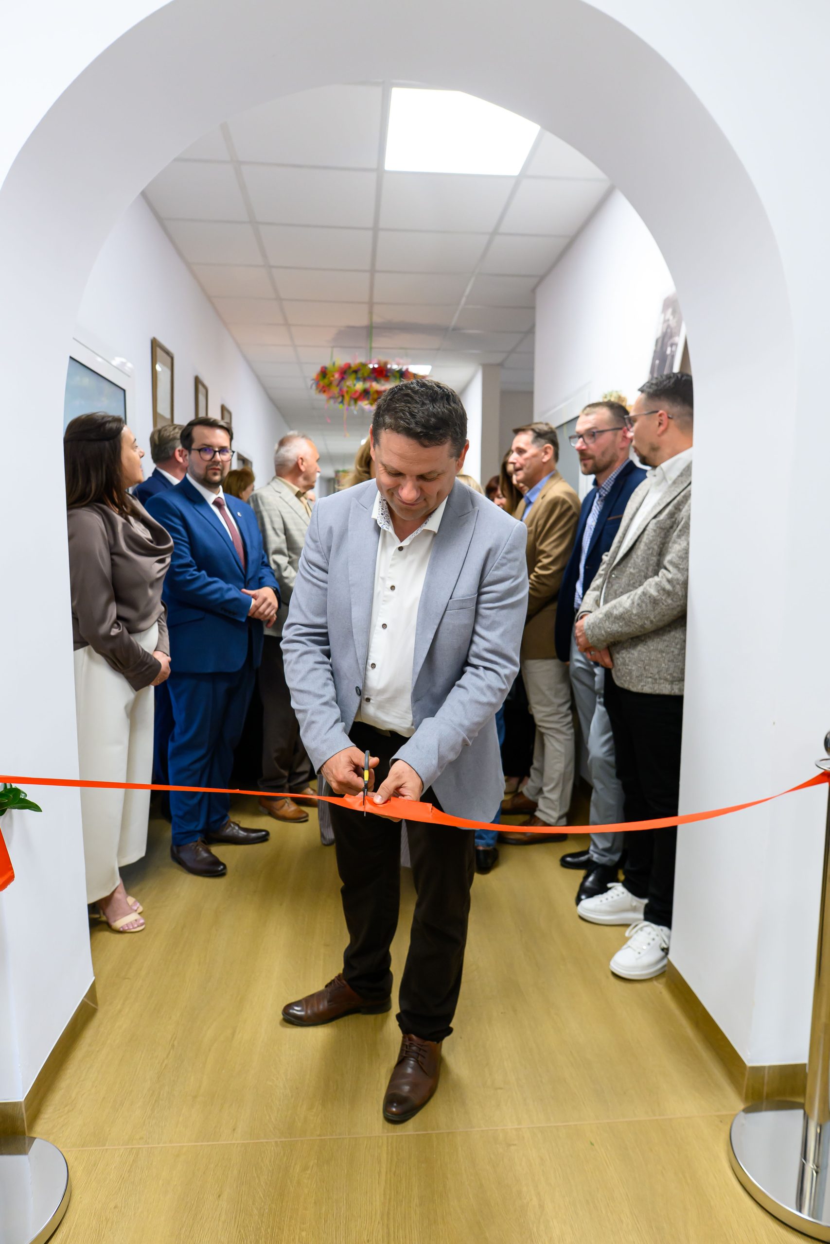 Osoba w jasnej marynarce przecina czerwoną wstęgę w korytarzu. W tle stoją elegancko ubrani ludzie. Jasne ściany i drewniana podłoga.