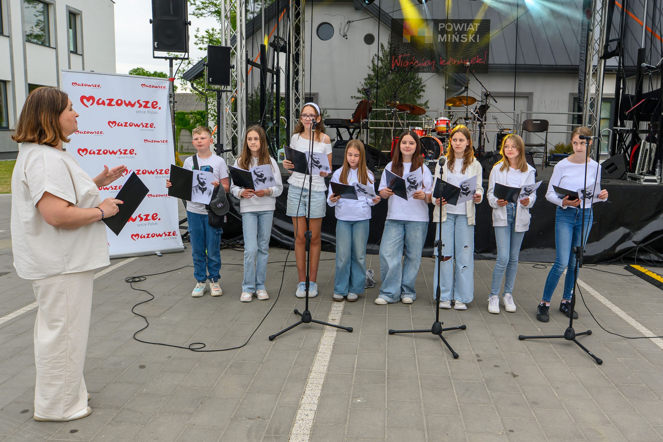 Grupa dzieci w białych koszulkach śpiewa na zewnątrz. Tło to scena z perkusją. Obok stoi dorosła osoba w kremowym stroju.