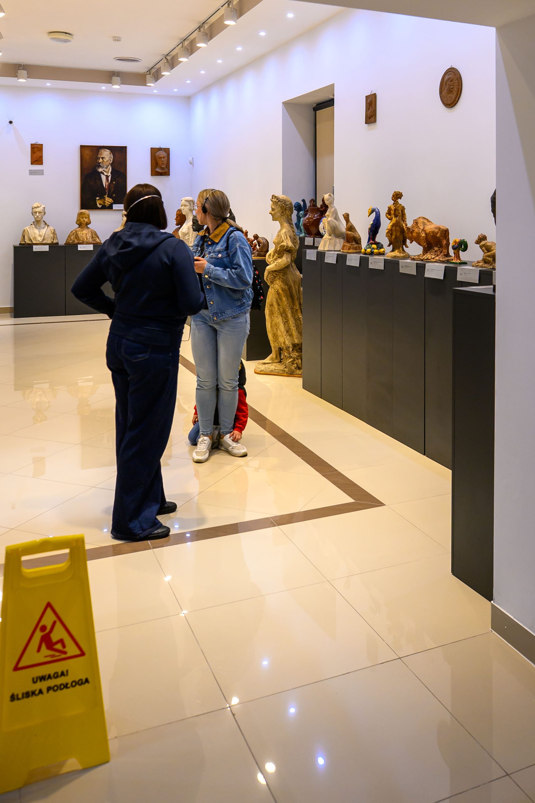 W galerii sztuki na jasnej podłodze stoją rzeźby i obrazy. Dwie osoby rozmawiają, a obok znak śliska podłoga. Ściany są białe, a rzeźby wystawione na ciemnych cokołach.
