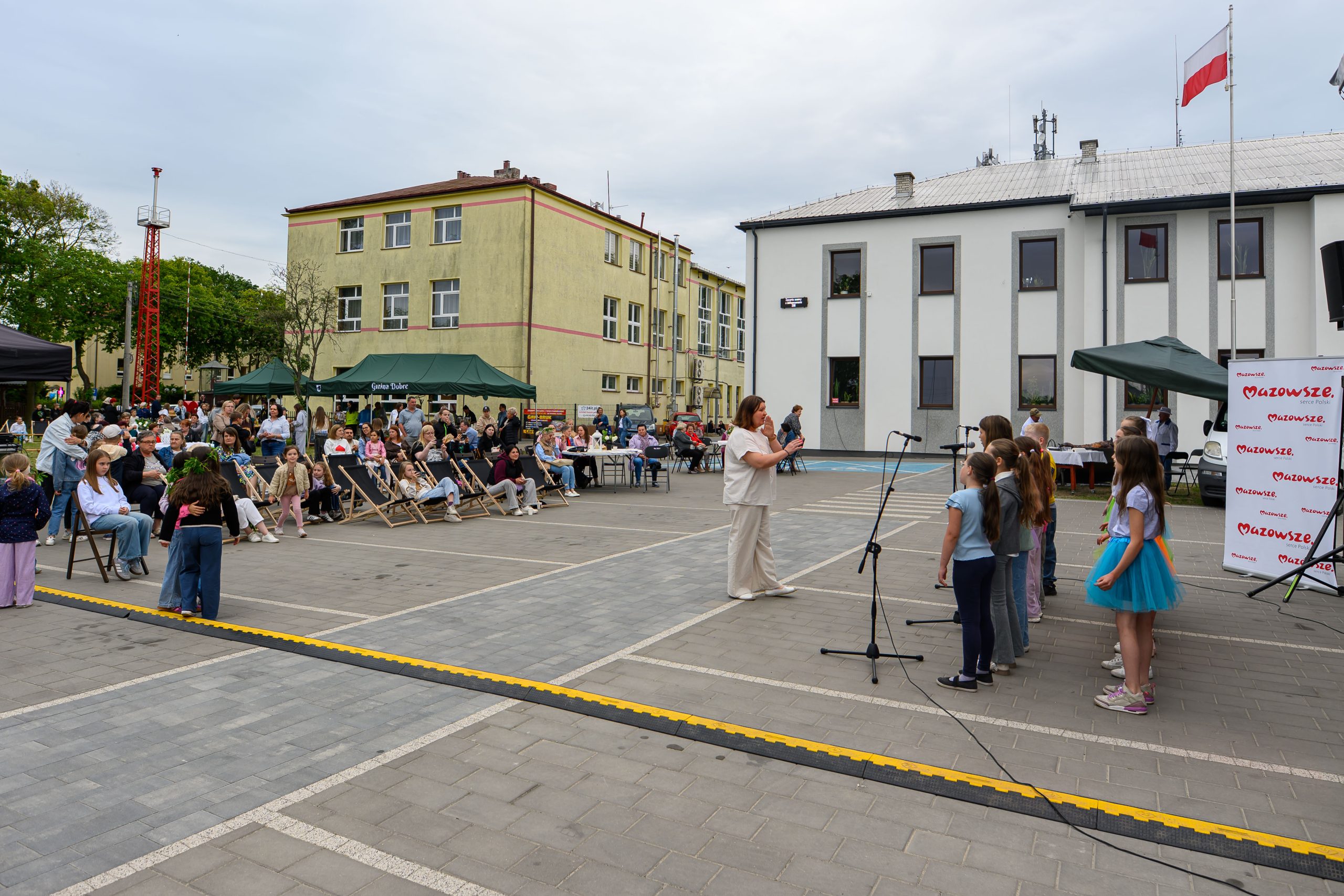 Na zdjęciu grupa dzieci na scenie, ubrana kolorowo, śpiewa dla publiczności na szarym placu z budynkami w tle.